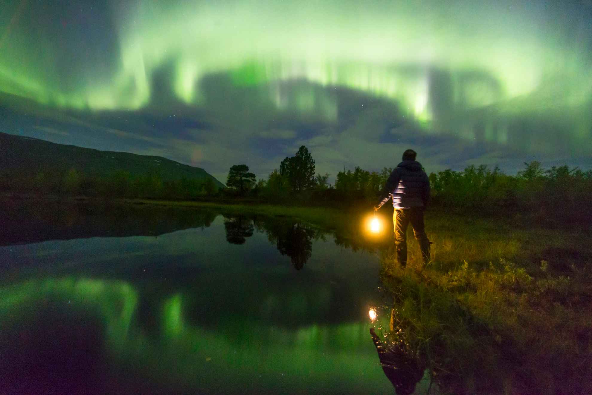A person holding a light looks at the green northern lights in the sky. The northern lights are reflecting in the lake next to the person.