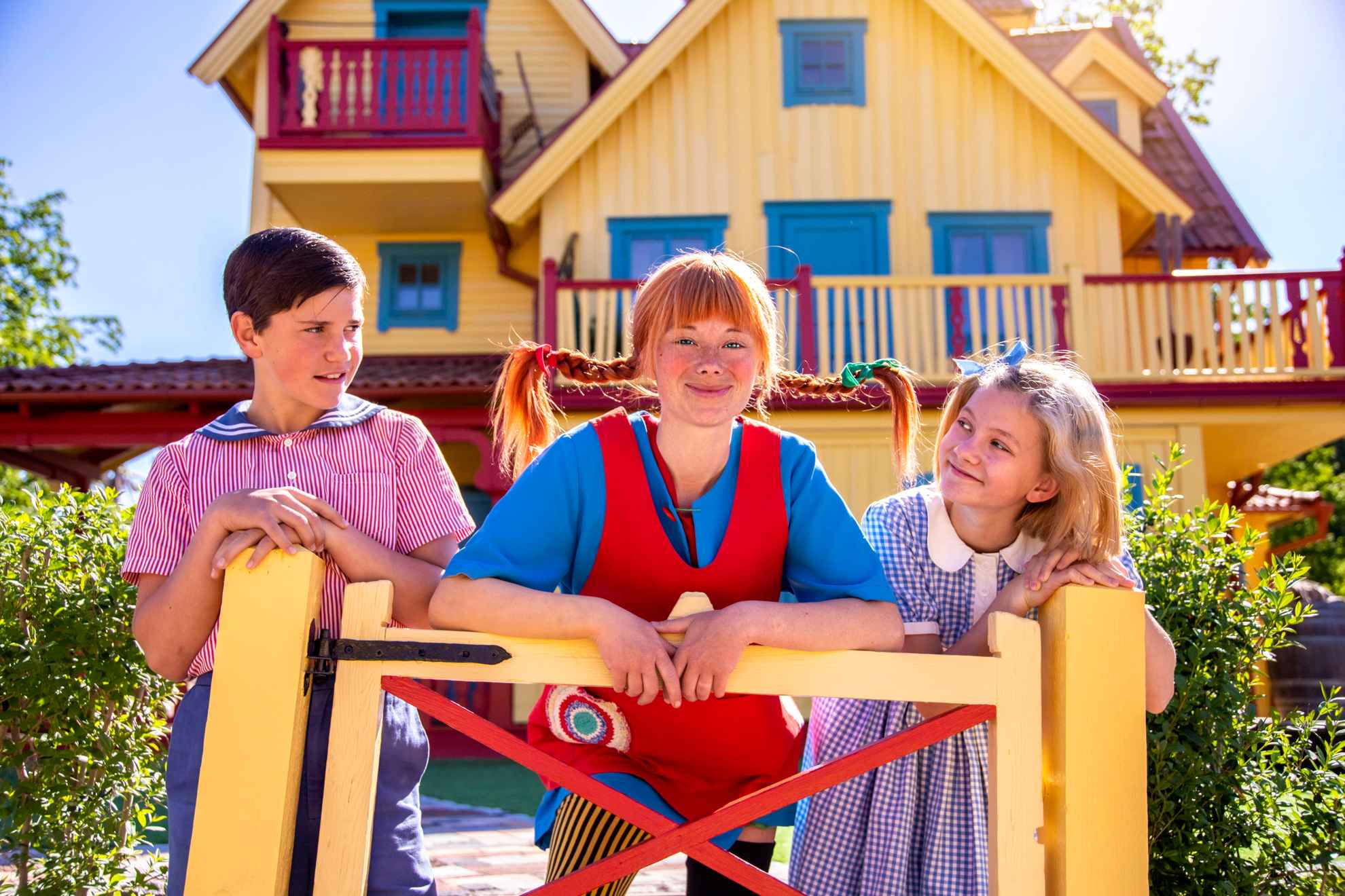 Actors dressed as the characters Tommy, Pippi and Annika stand by a yellow gate in front of a yellow house (Villa Villekulla)..