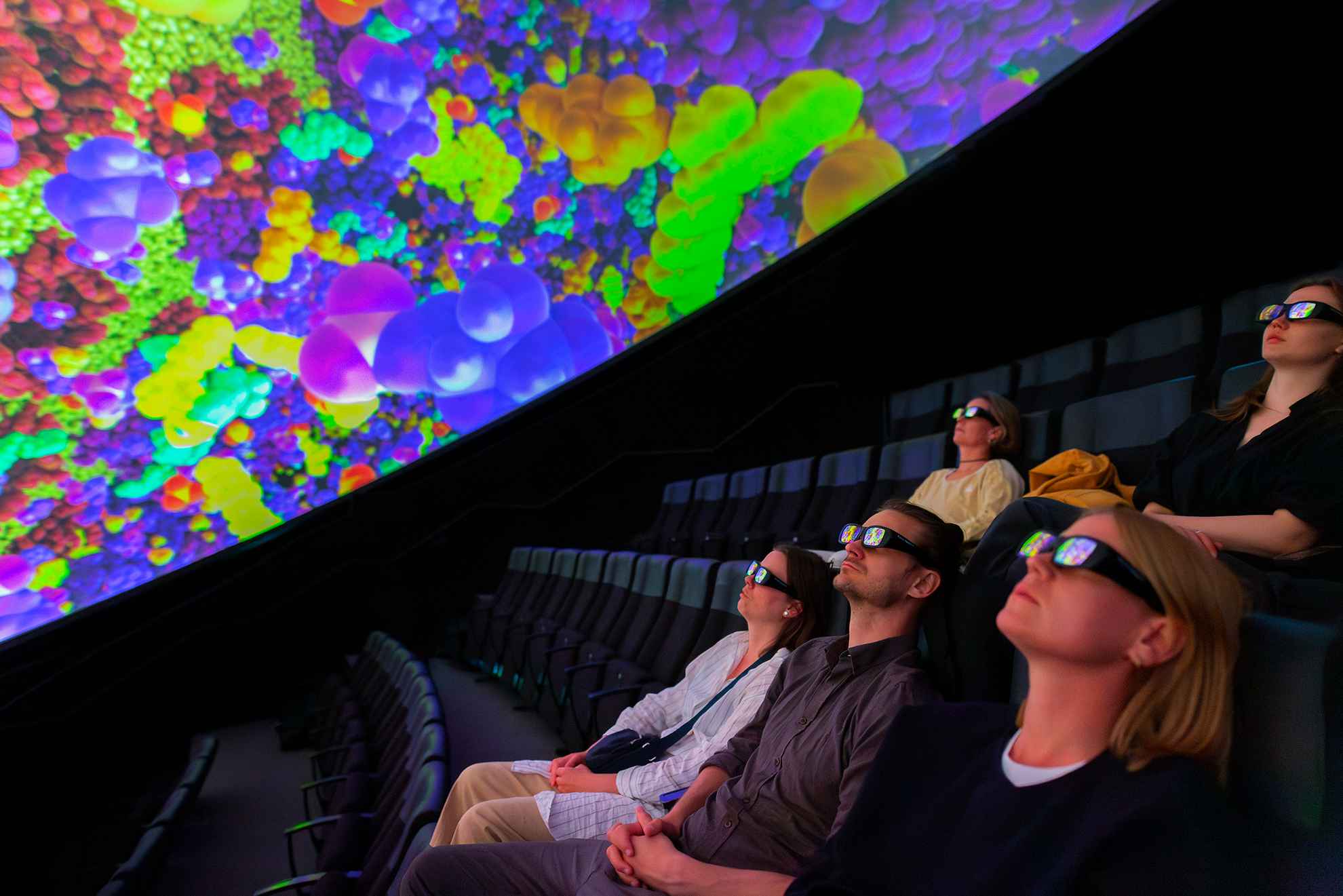 A group of people with 3D glasses sit in chairs and watch the screen in the Wisdome Stockholm.