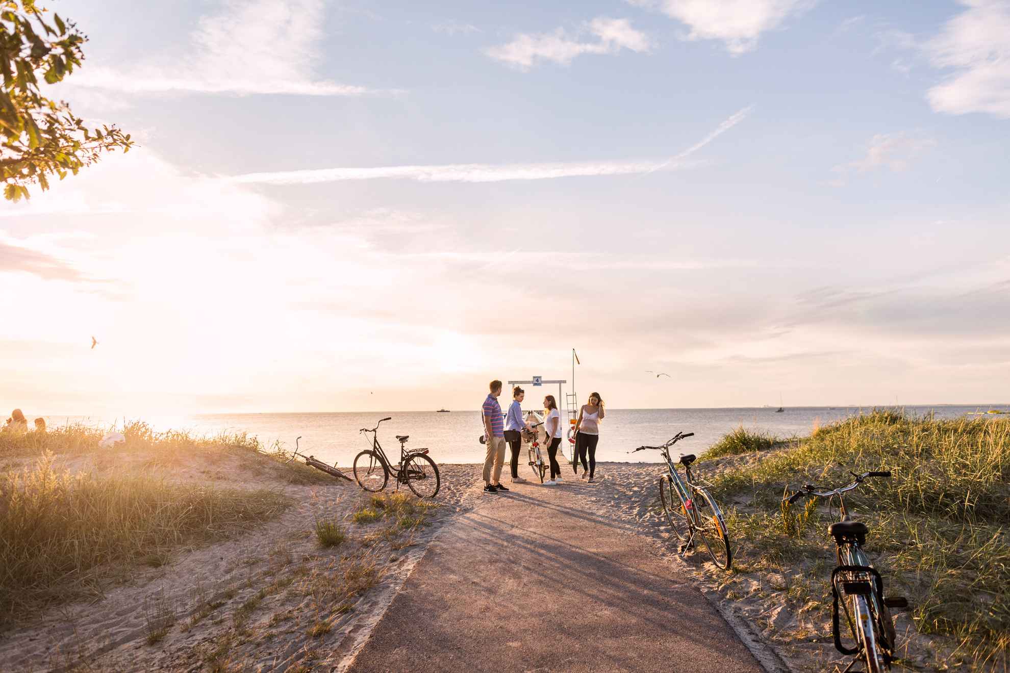 Sunset at Ribersborg beach, a man and three women is seen at a bathing pier at the beach. There are server bikes parked along the path to the beach.
