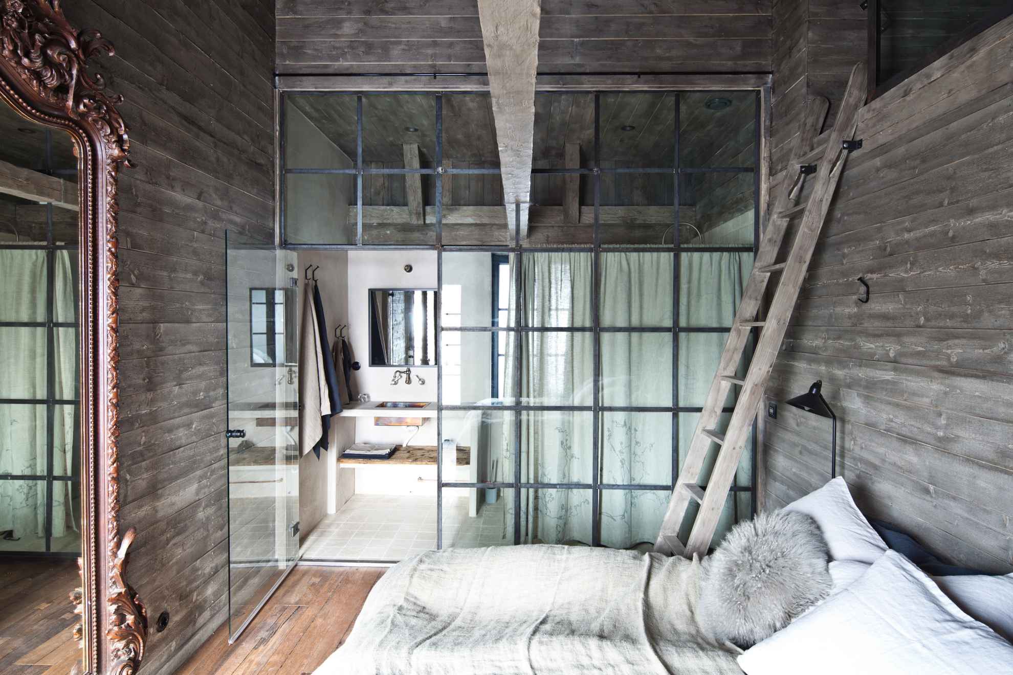 A hotel room with wooden walls. The room has a glassed-in bathroom with a curtain. There is also a ladder leading to a sleeping loft.