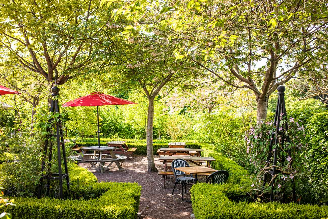 Café tables surrounded by lush trees and well-trimmed shrubs. A red parasol stands by one of the tables.