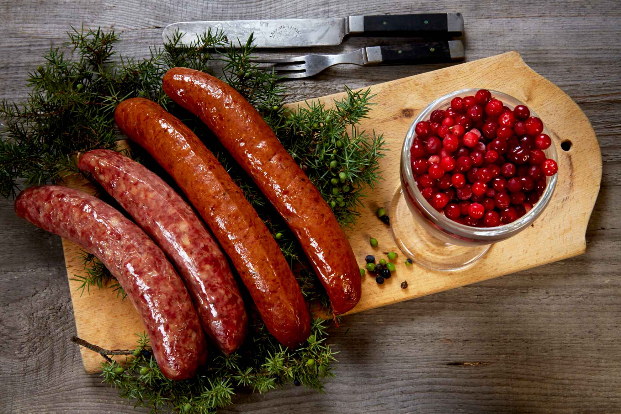 Four sausages of lard on a bed of juniper twigs on a wooden cutting board together with a bowl of lingonberries.