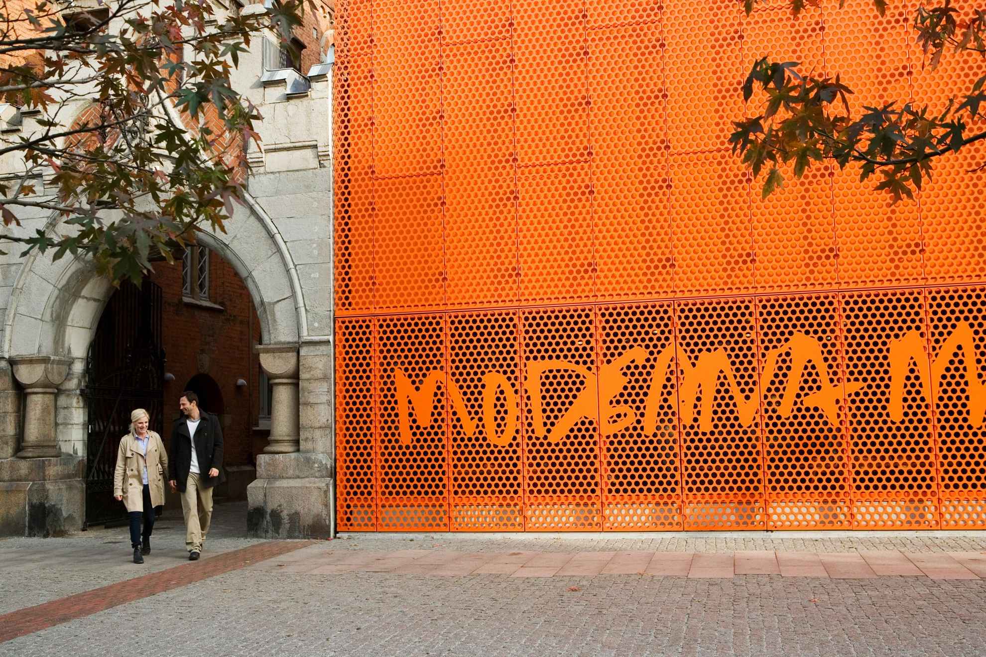 A man and a woman are walking outside and past the orange sign of the Moderna Museet in Malmö.