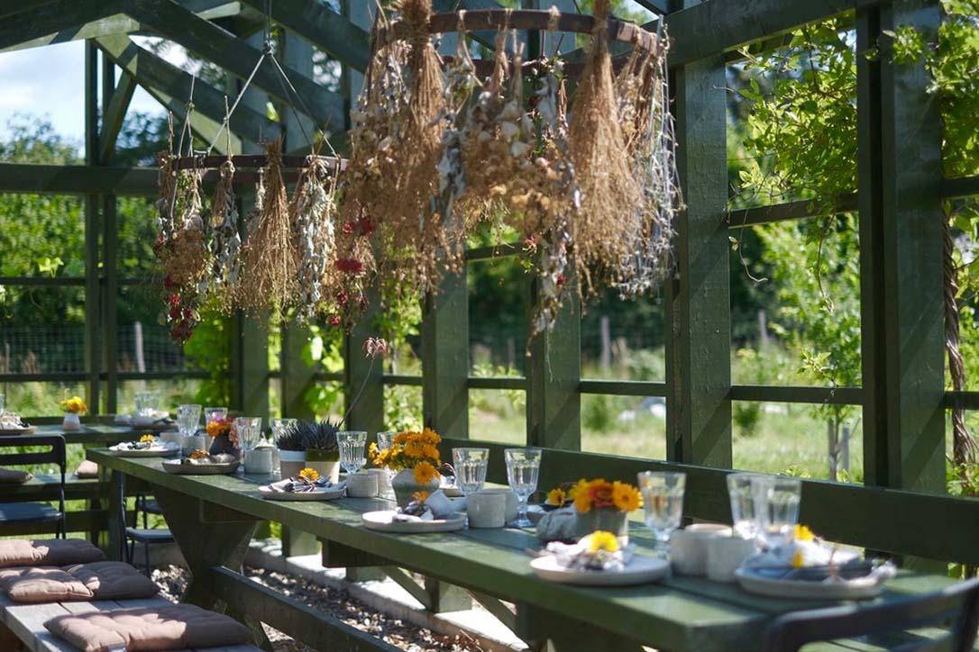 A green long wooden table is set with glasses, plates and yellow flowers. The table stands outdoors in a wooden building.