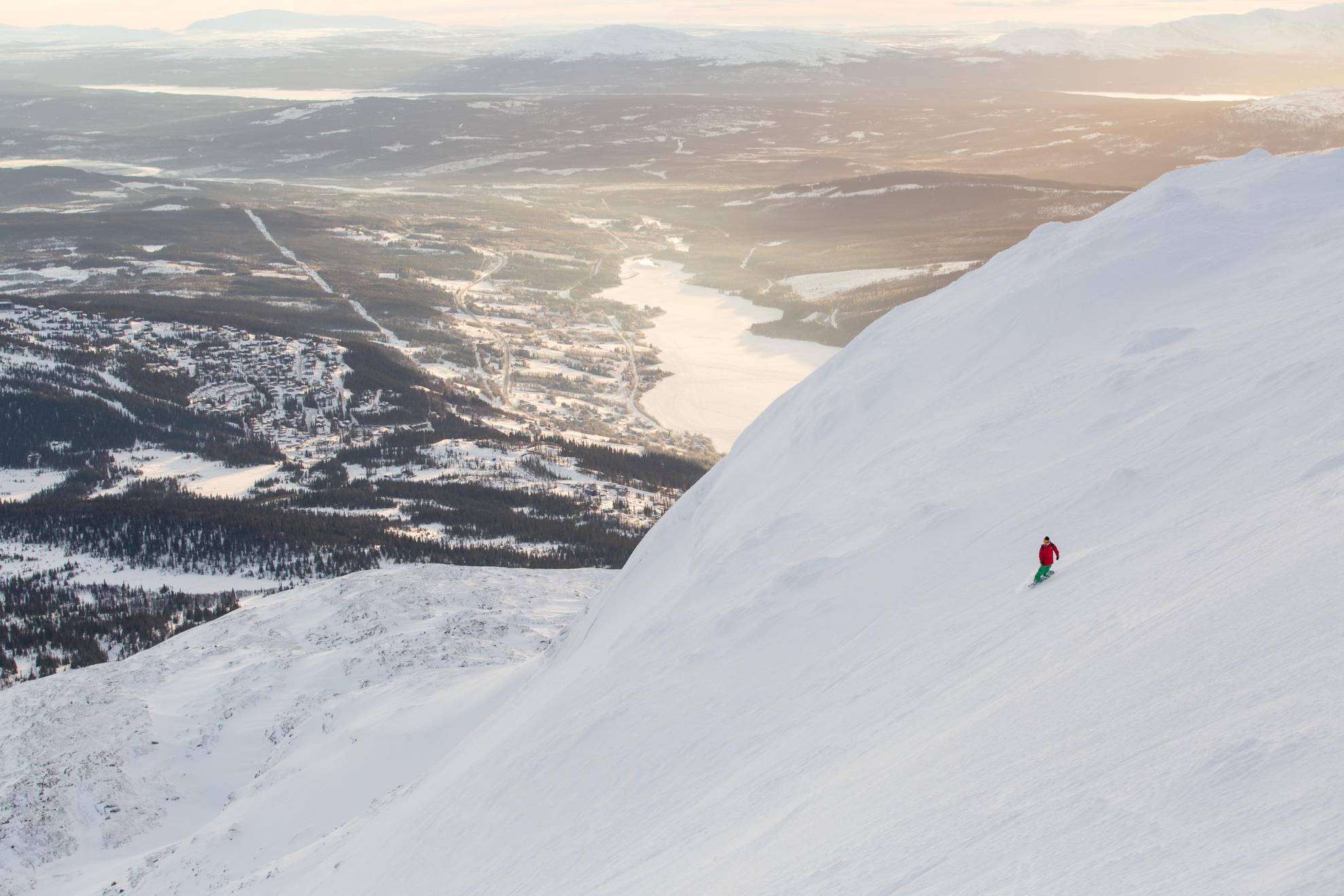 A scenic view of the Åre valley and a river seen from one of the peaks. A person skis down the slope.