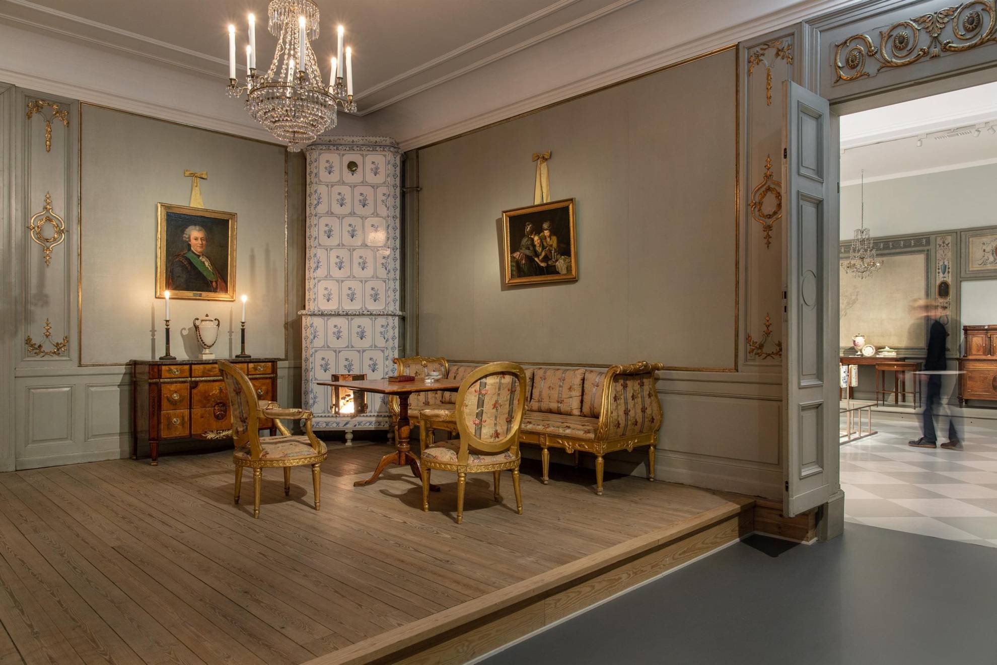 18th-Century furniture at an exhibition. A tile stove, a sofa and two chairs, a table and a chest of drawers, a crystal chandelier hanging from the ceiling and two paintings on the wall.