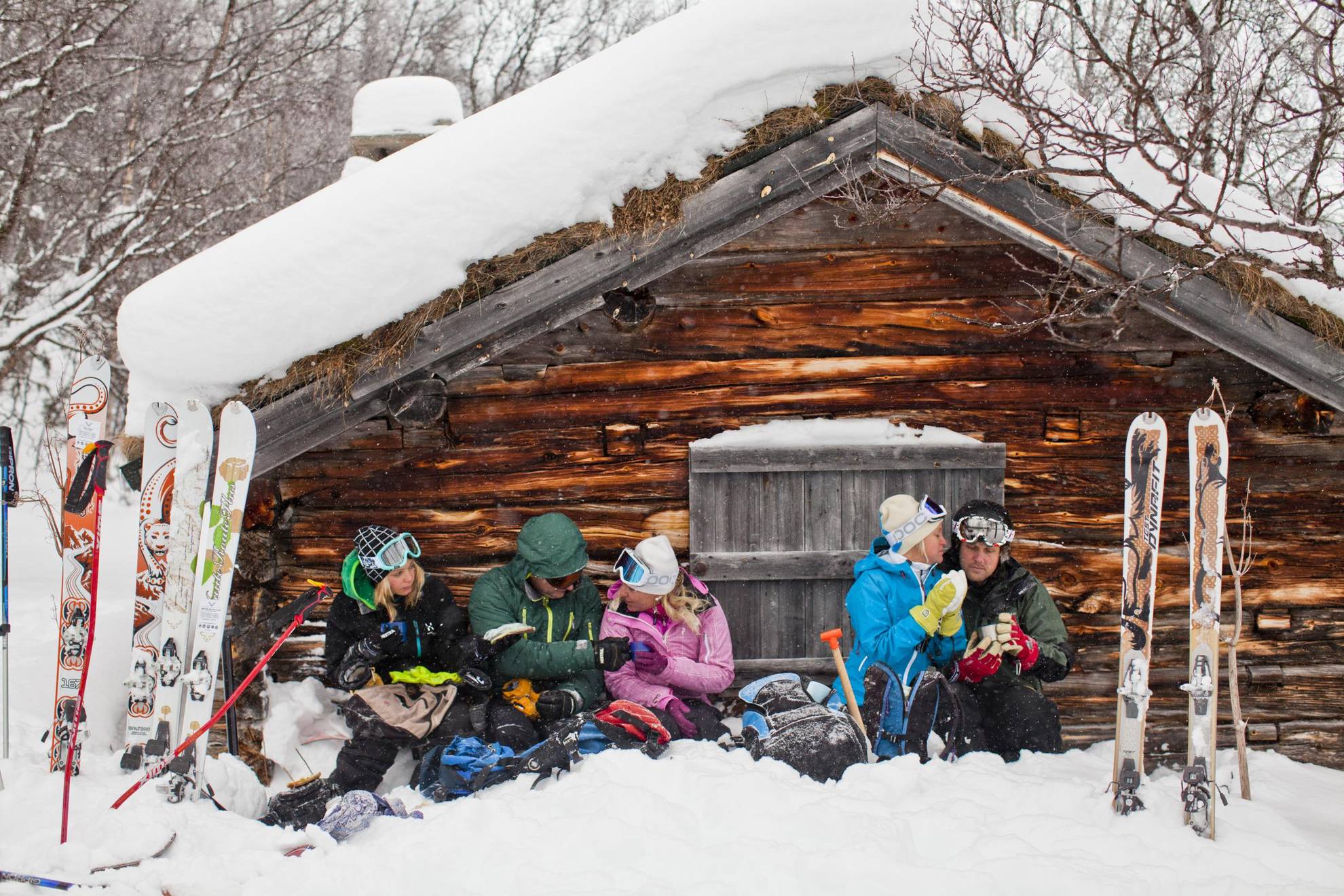 Five people are sitting in the snow in front of a wooden house in the mountains and having a coffee break from skiing.