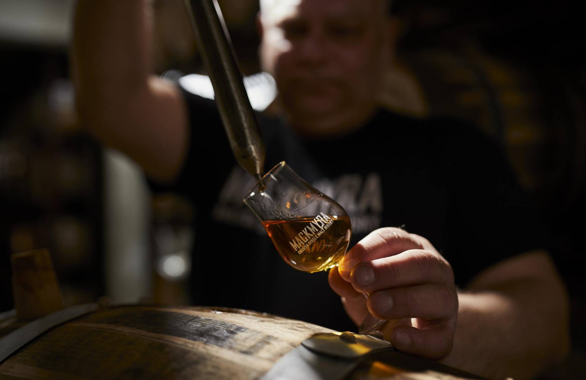 Whisky is poured from a pipette into a whisky glass with the text Mackmyra on it. An oak barrel is in the foreground.
