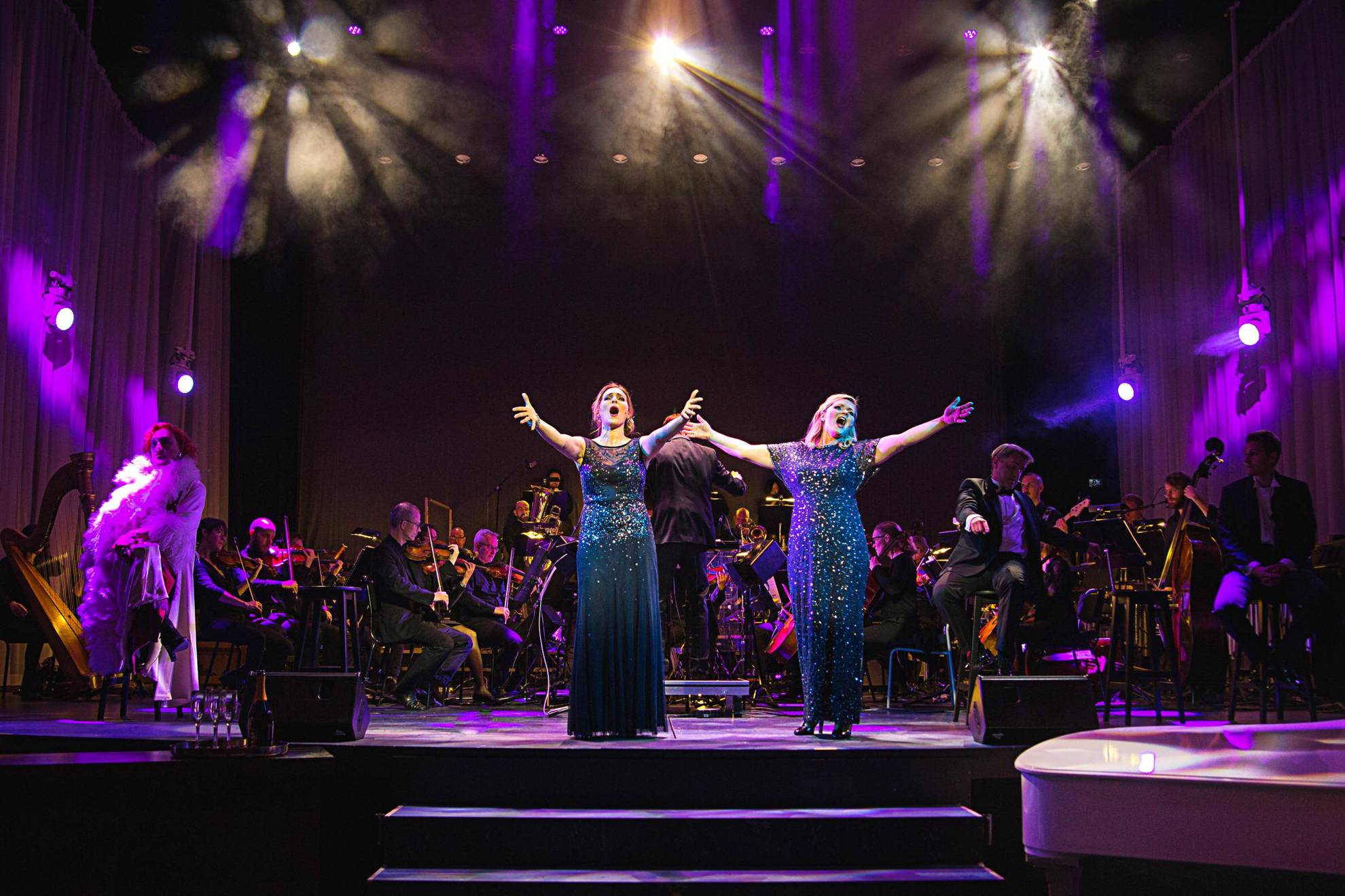 Two women in blue sparkly dresses sing on a stage during a performance. Behind them is an orchestra.