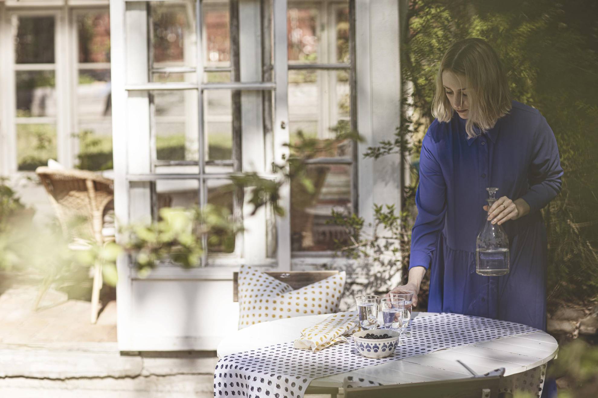A woman puts glass of water on a table that is set with a table cloth and a bowl of blueberries. In the background you see the doors to a house and there is a branch blurry in the front.
