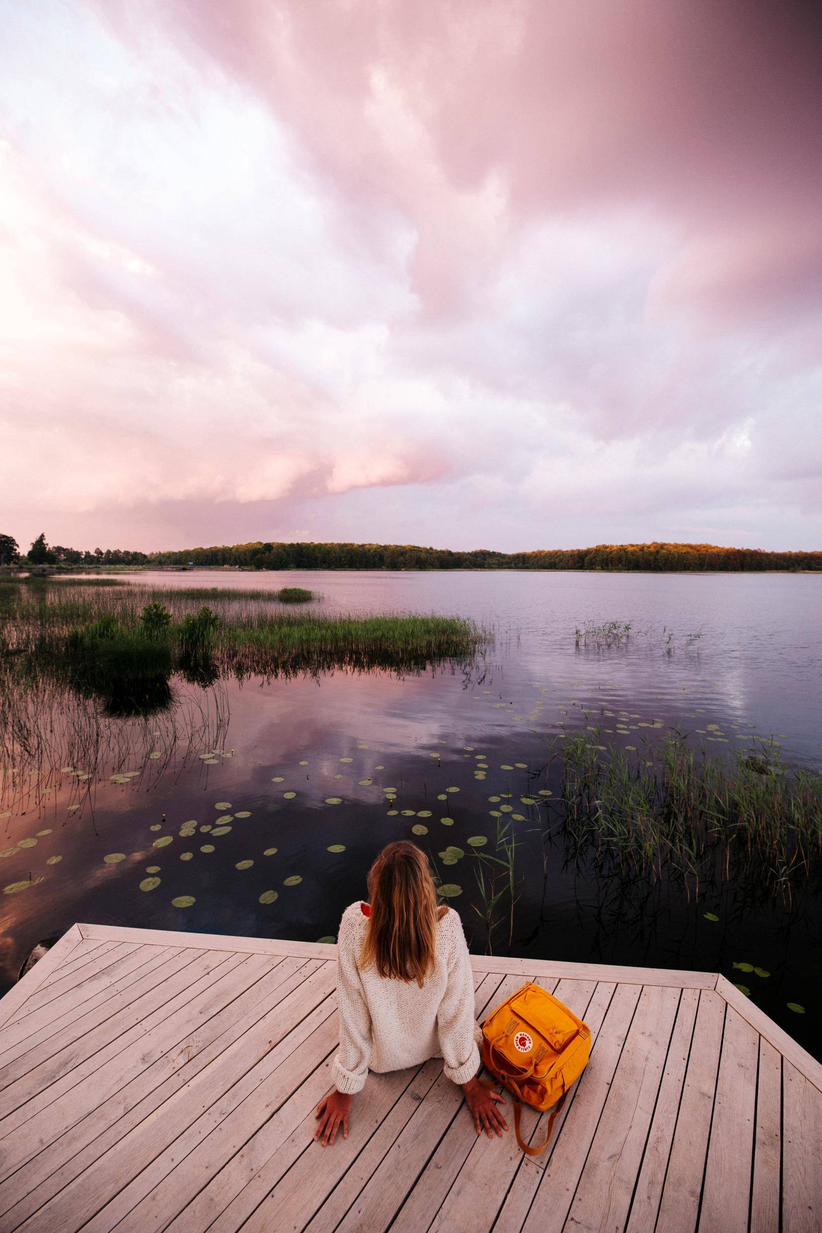 A woman sitting back on a boardwalk by a lake, looking out over a lake which is surrounded by greenery.