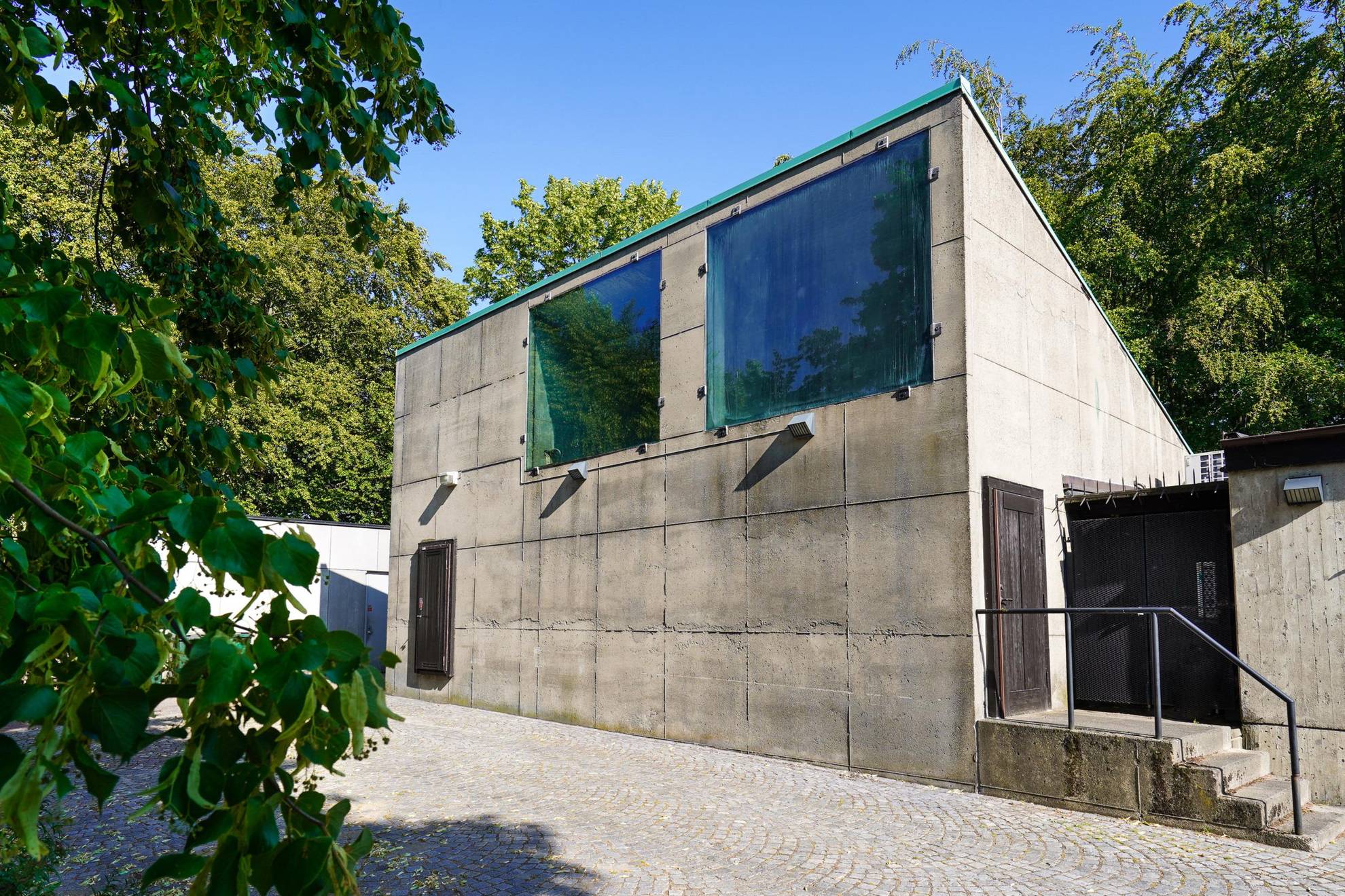 The house called 'The flower kiosk' is a simple concrete house where the windows are attached with black sealant. It is surrounded by greenery.