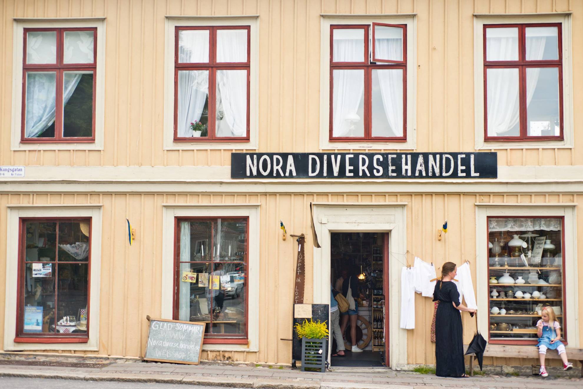 A small shop in a yellow wooden house on a street in a old town. A sign above the entrance says Nora Diversehandel. A woman is standing  and a girl is sitting on a bench in front of the house.