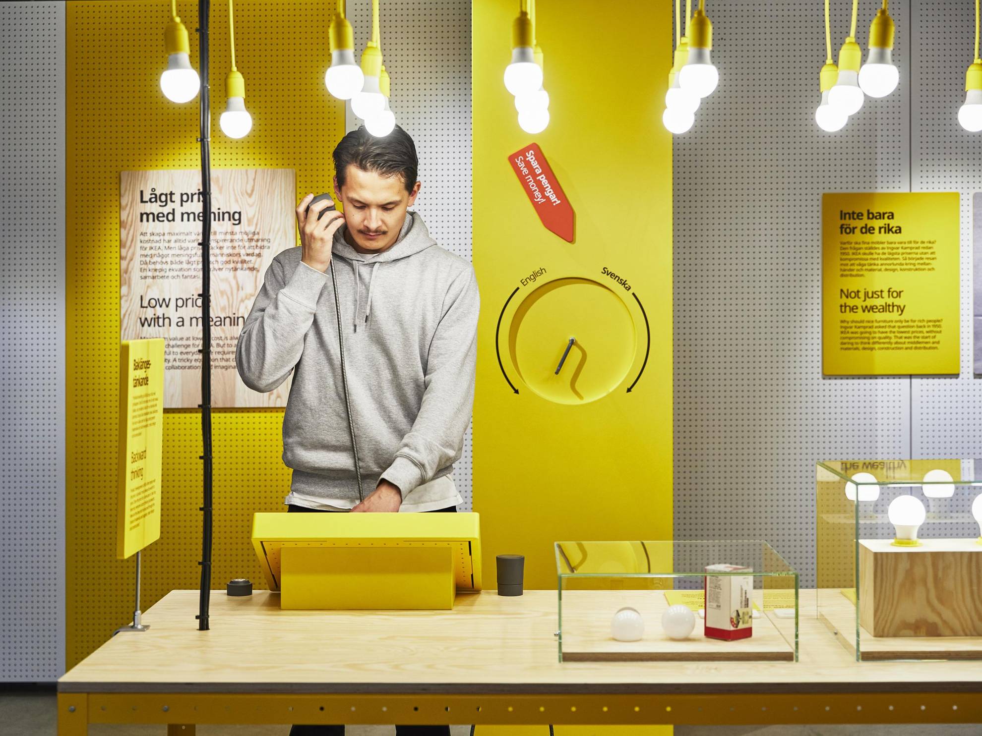 A man is standing by a table looking at a yellow display in one of Ikea's exhibitions. There are light bulbs hanging from the ceiling and glass boxes with light bulbs in them on the table.