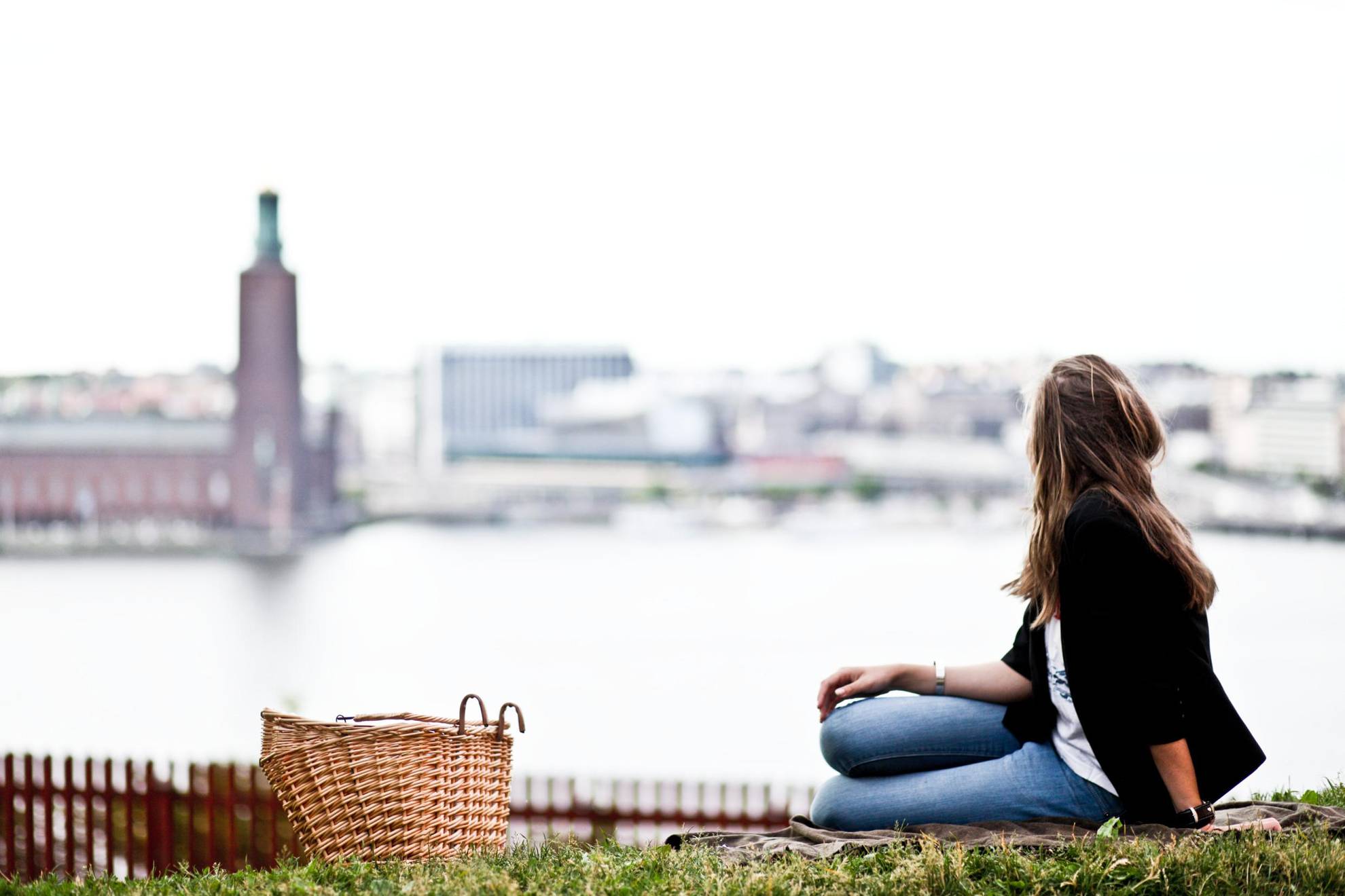 A woman with a weaved basket sits in a park overlooking Lake Mälaren and Stockholm City Hall.