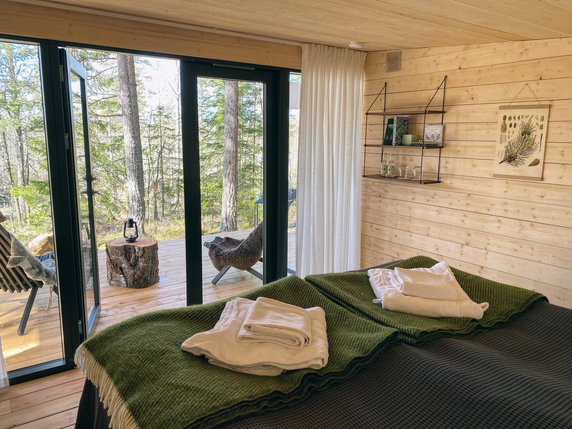 Two beds are in a room with wooden walls and floor. The room has glass doors to a patio with two chairs overlooking nature.