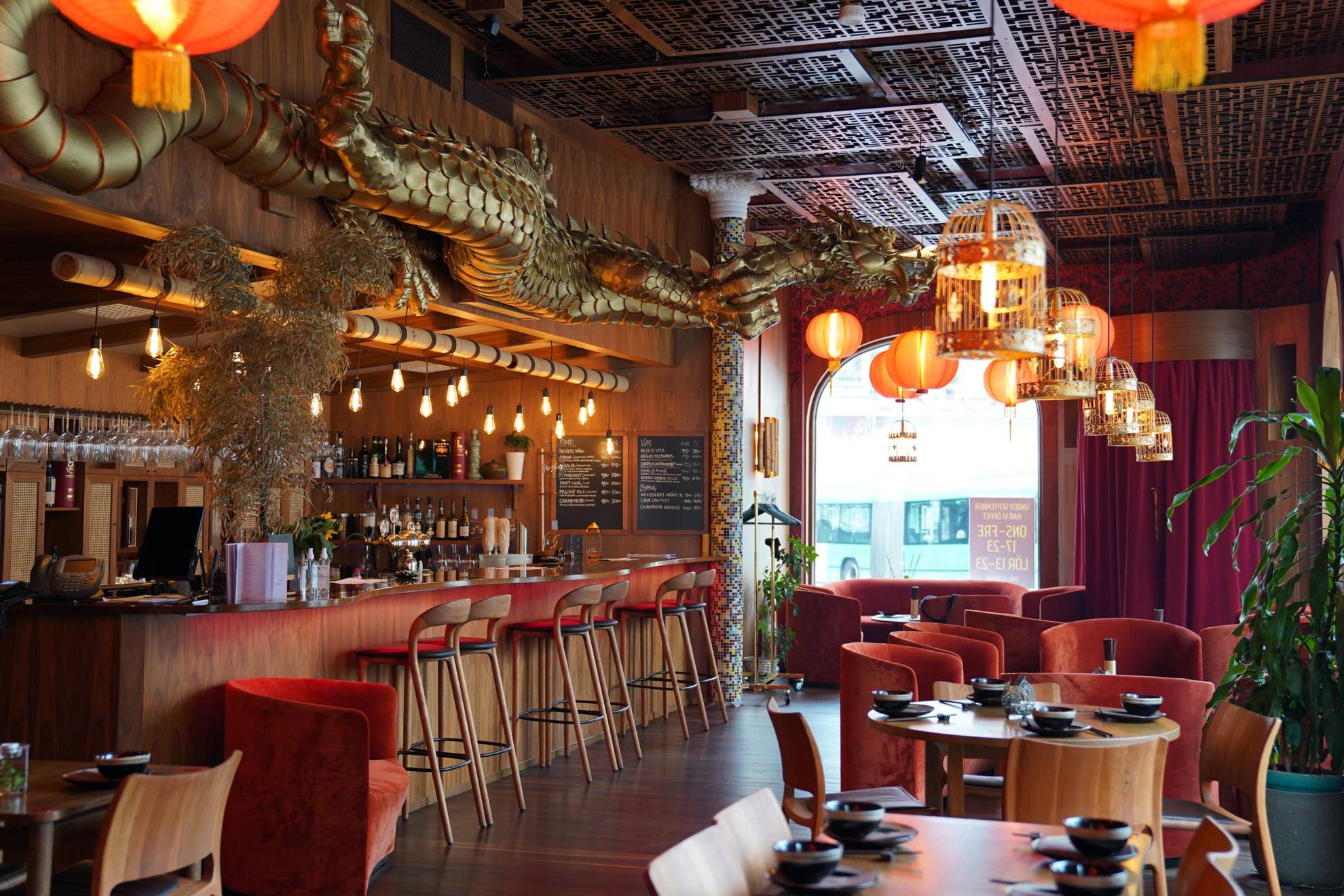 Inside a Chinese restaurant. To the left a bar with six bar stools. A large golden dragon is placed above the bar. To the right tables and chairs. The tables are set with plates, small bowls and chop sticks. Orange lamps are hanging from the ceiling.