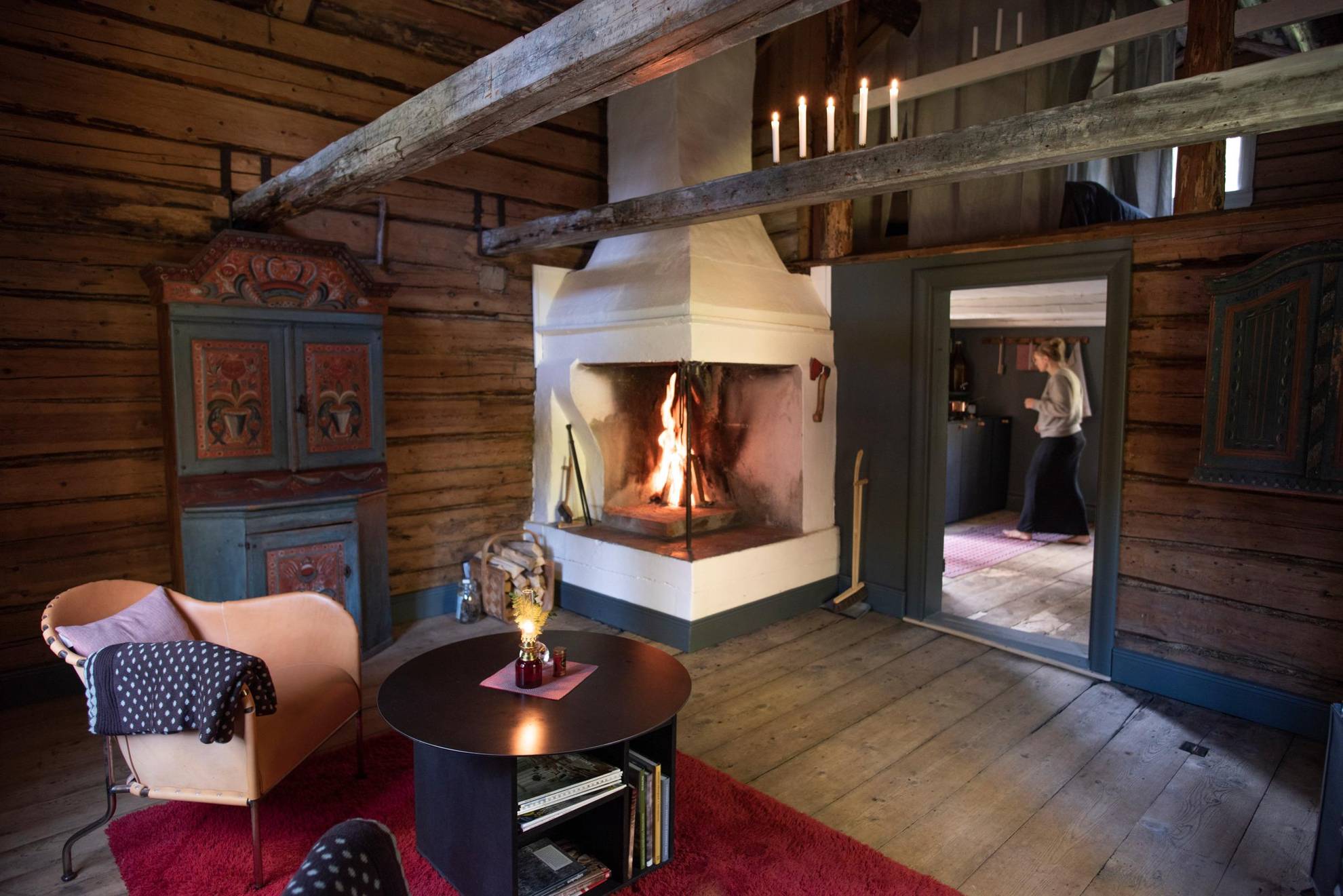 A fire burns in the hearth of an old wood cabin furnished with typical Hälsingland design as well as modern furniture, as you see a woman walking through the kitchen in the background.