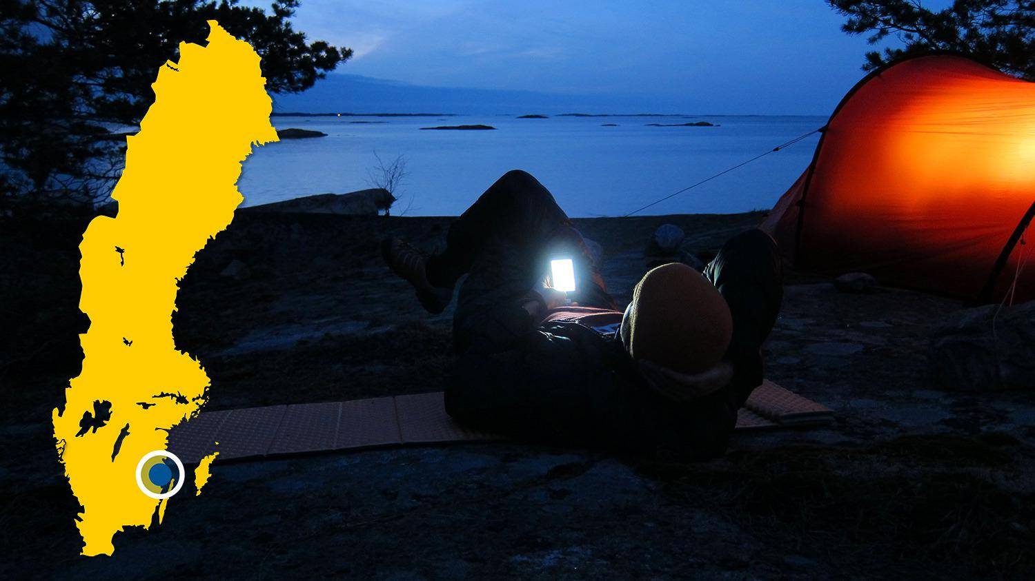 A tent is set up on a beach near the water. It is night and a person is lying next to the tent looking at his phone.