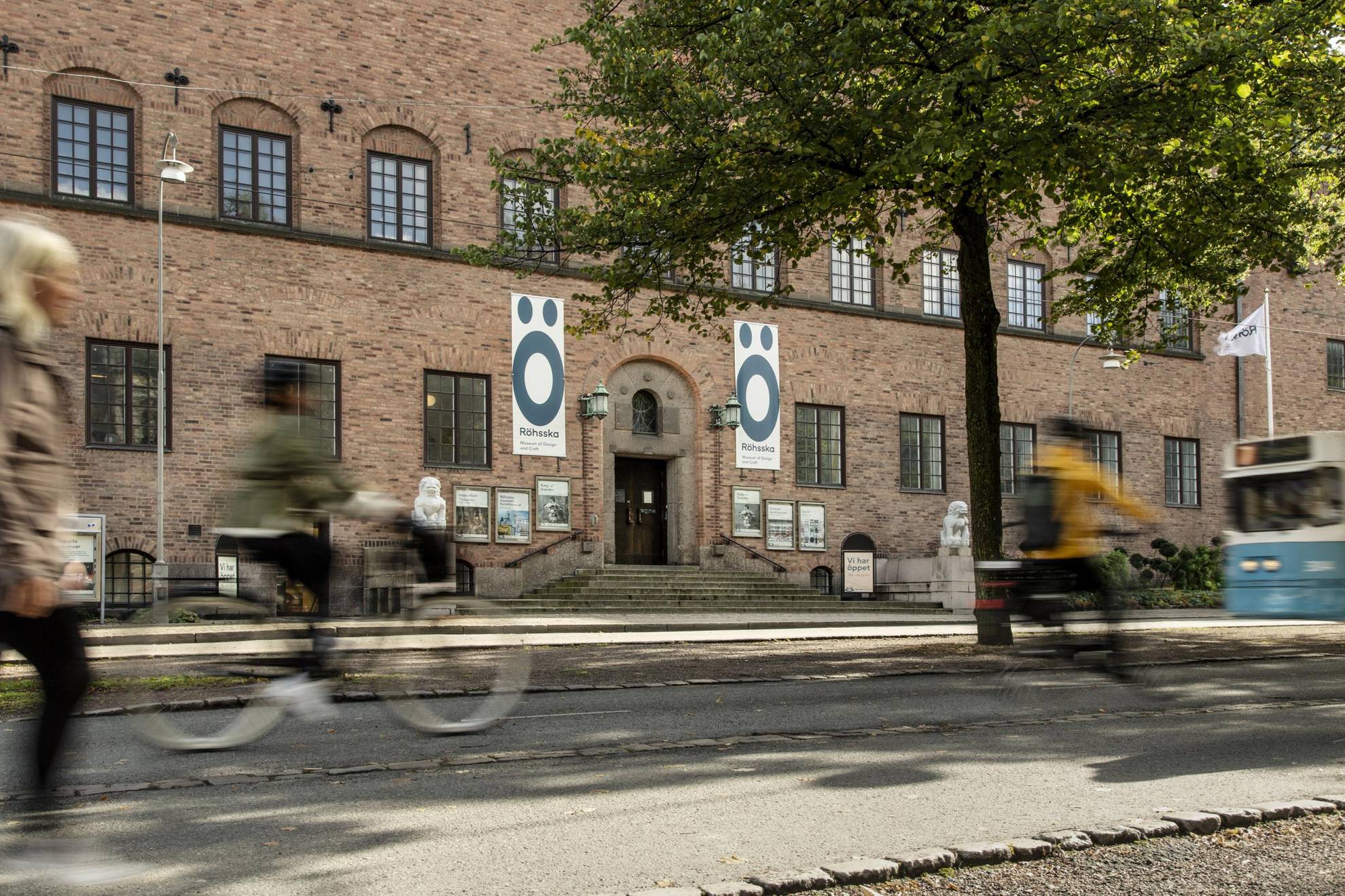 Two biking and one walking person are seen blurry passing the entrance to and the facade of Röhsska museum in Gothenburg. The building is made of bricks and on each side of the entrance there is white stone statues with figures looking a bit like lions.