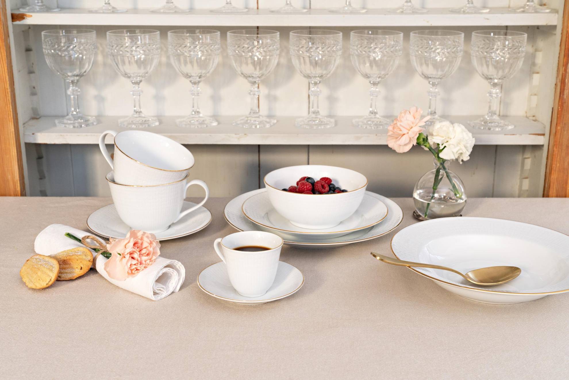 A tableware service called “Swedish Grace” on a table. Plates in different sizes a bowl with berries, two tea cups and a cup of coffee. Behind there are a shelf with several wine glasses.