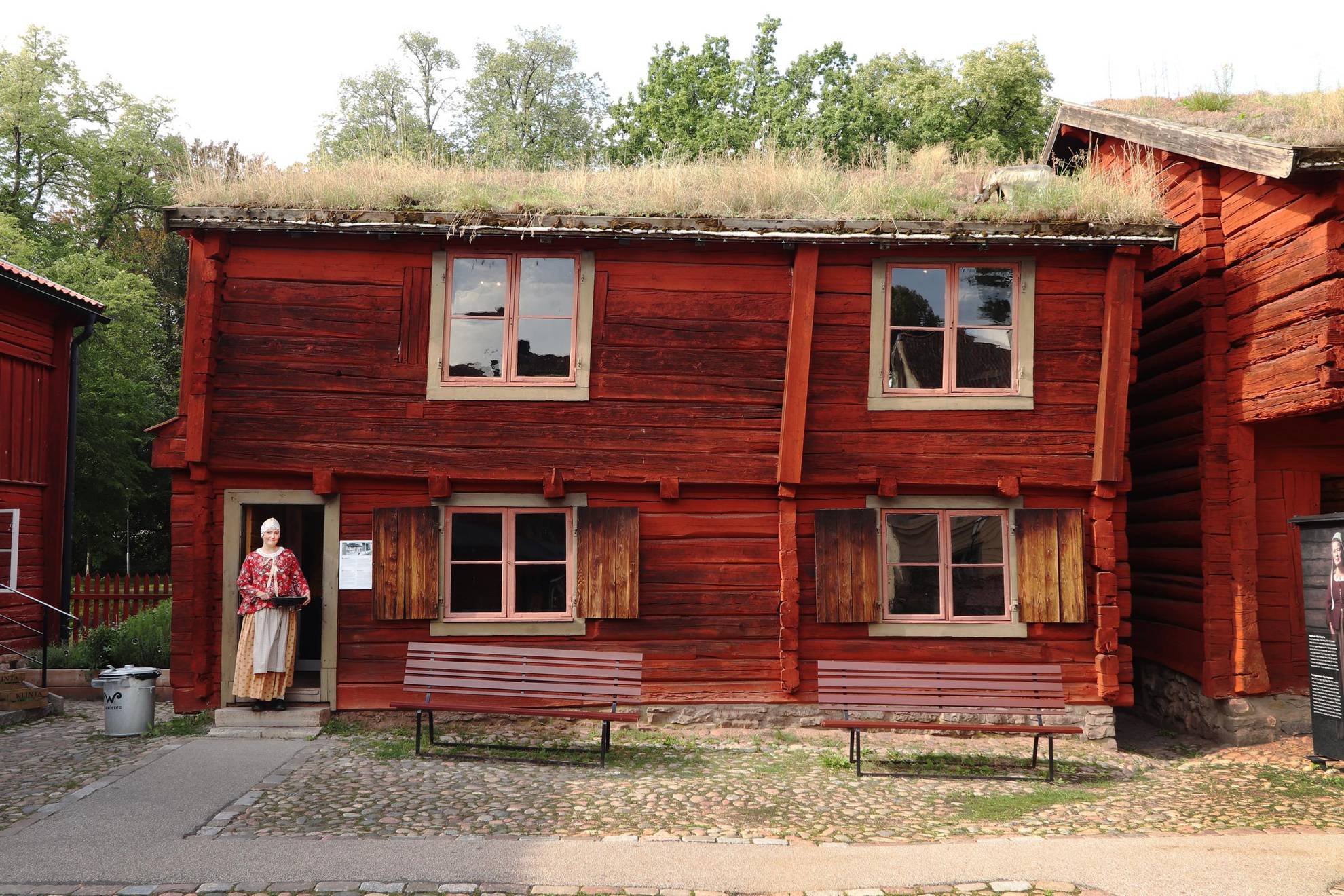 An old red painted wooden house with a sod roof. A women is standing at the entrance of the house.