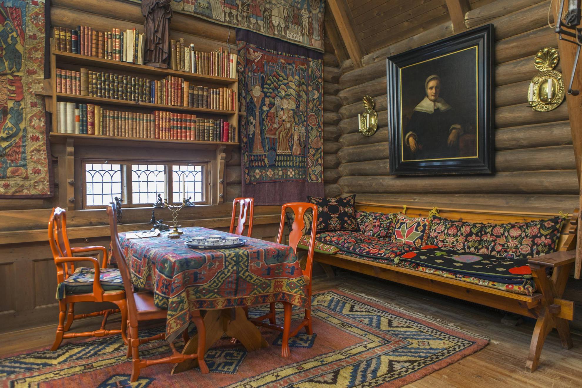 The colourful dining hall of Anders Zorn, with woven tapestries, bright red chairs around a table with a woven cloth, a bookshelf and a wooden couch.