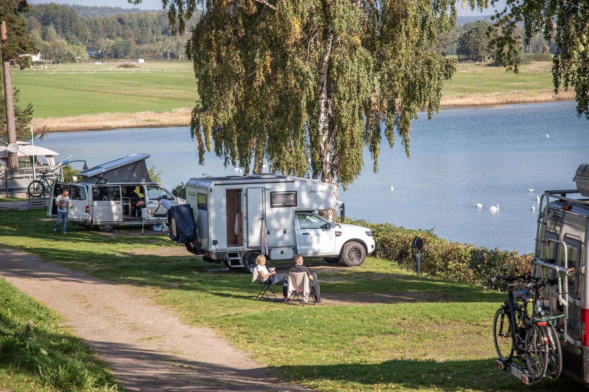 Two people are sitting in chairs outside their camper next to a lake.
