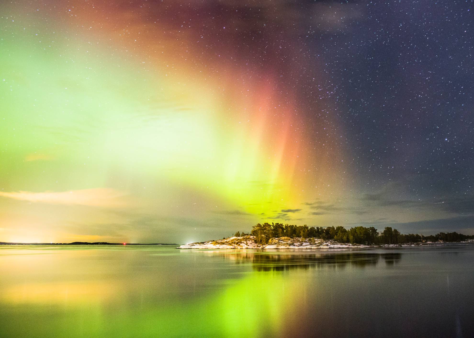 The northern lights in the sky are reflected in the water below. A city is visible from afar.