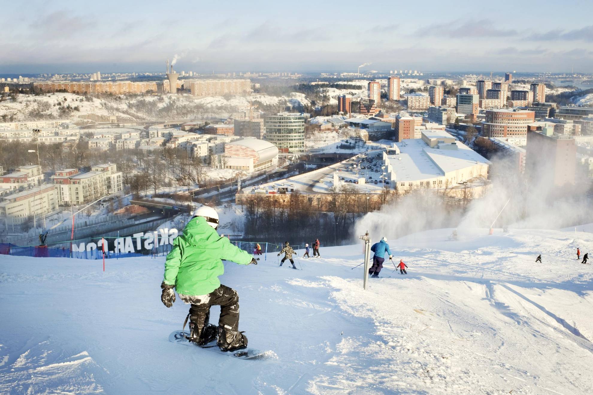 People are going down the slopes of Hammarbybacken on skis and snowboards, looking out over downtown Stockholm.