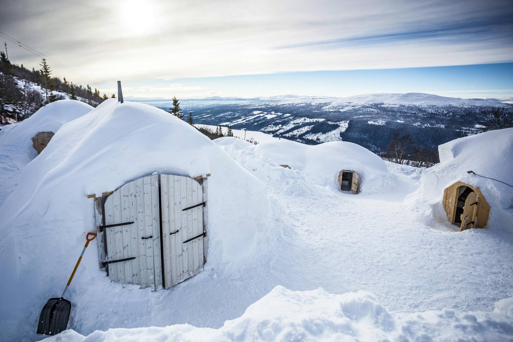 Igloos with small wooden doors are situated on a mountain. A shovel is placed against one of the igloos. In the background is a river, snow-covered mountains, and forest.