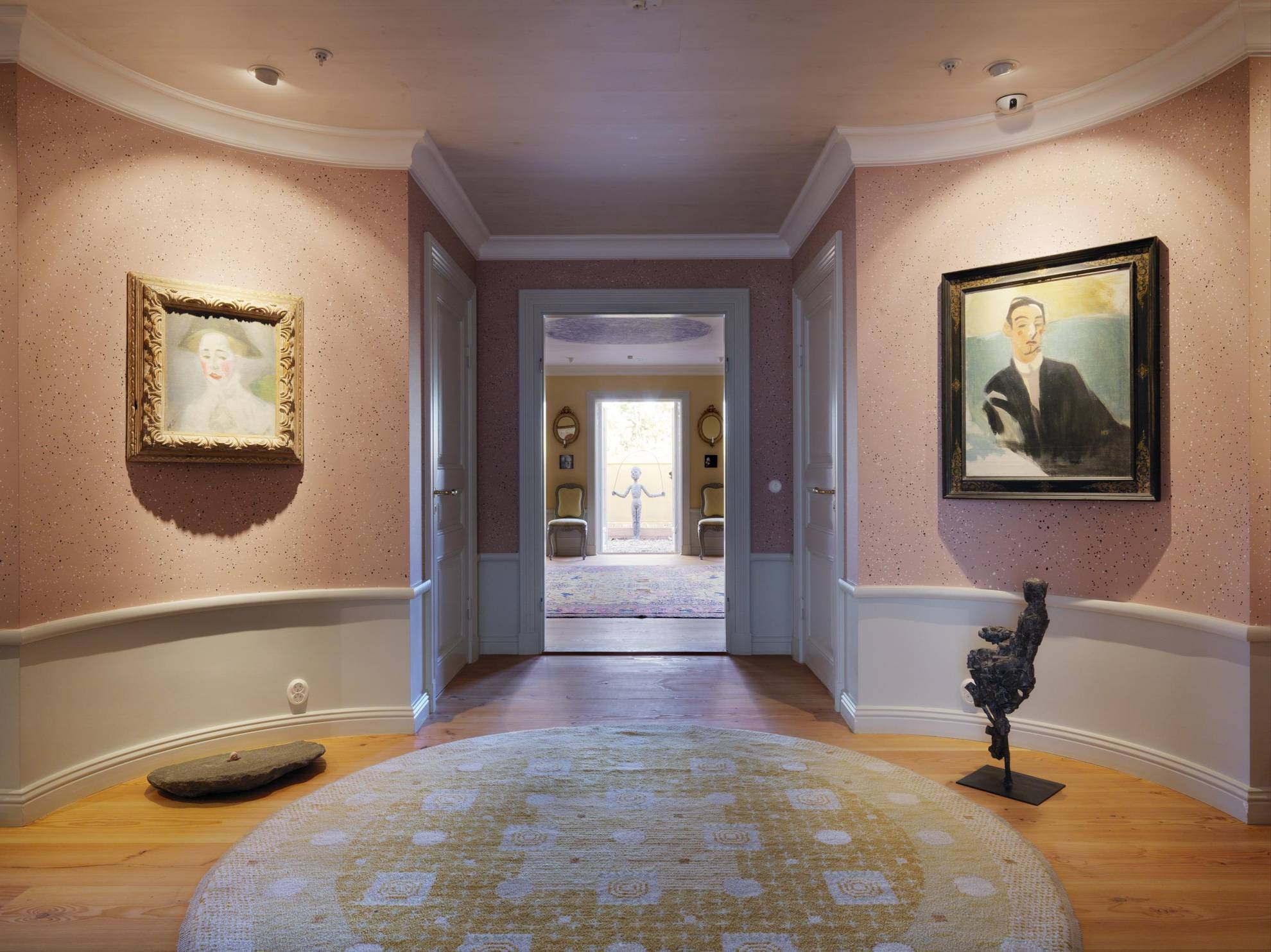 A hallway with pink tapestry and to paintings on each side of a doorway leading into another room.