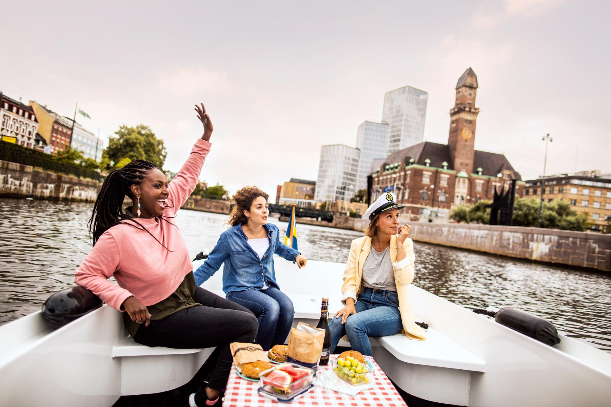 Three young happy women on an excursion in a boat on Malmö's canal with Malmö Live and the central station in the background.