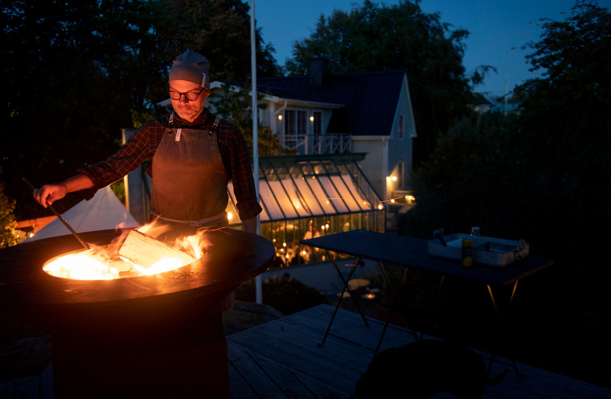 A man with a leather apron is standing by a fire during the evening.