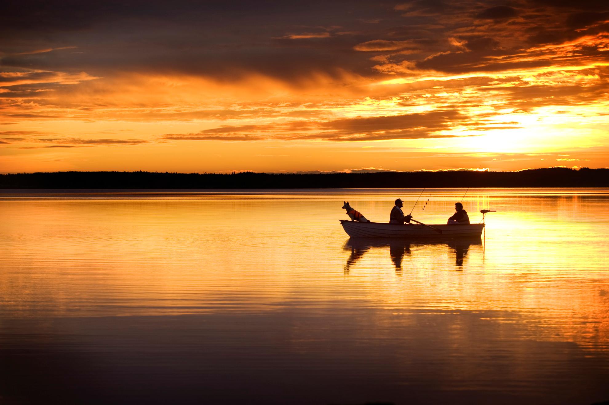 Two men and a dog in a small boat on a lake are fishing in the midnight sun. The orange sky is reflecting in the lake.