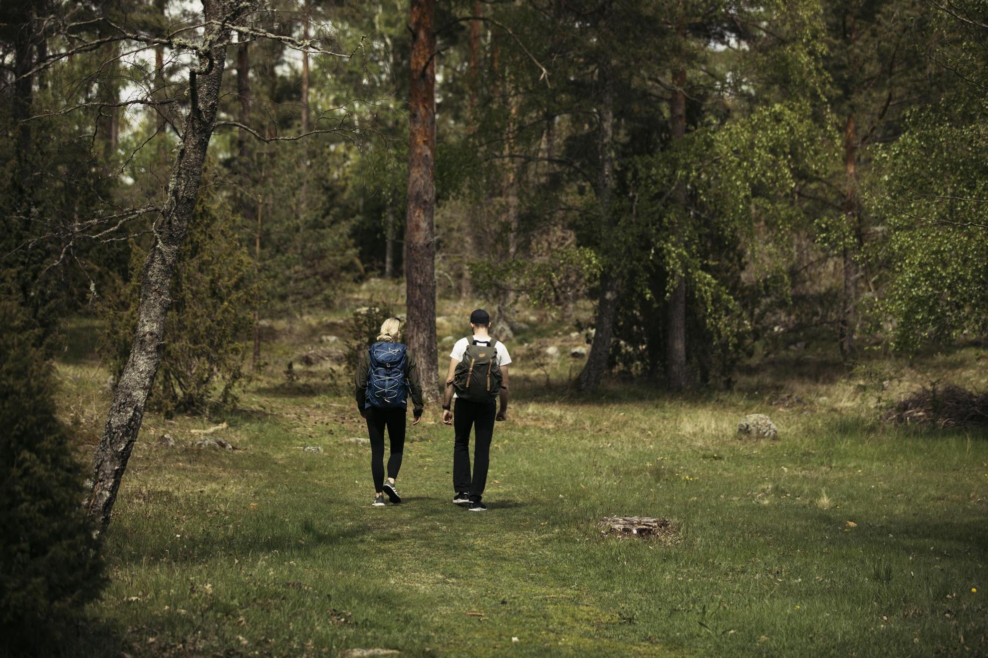 Two people with backpacks are walking in a meadow surrounded by greenery.