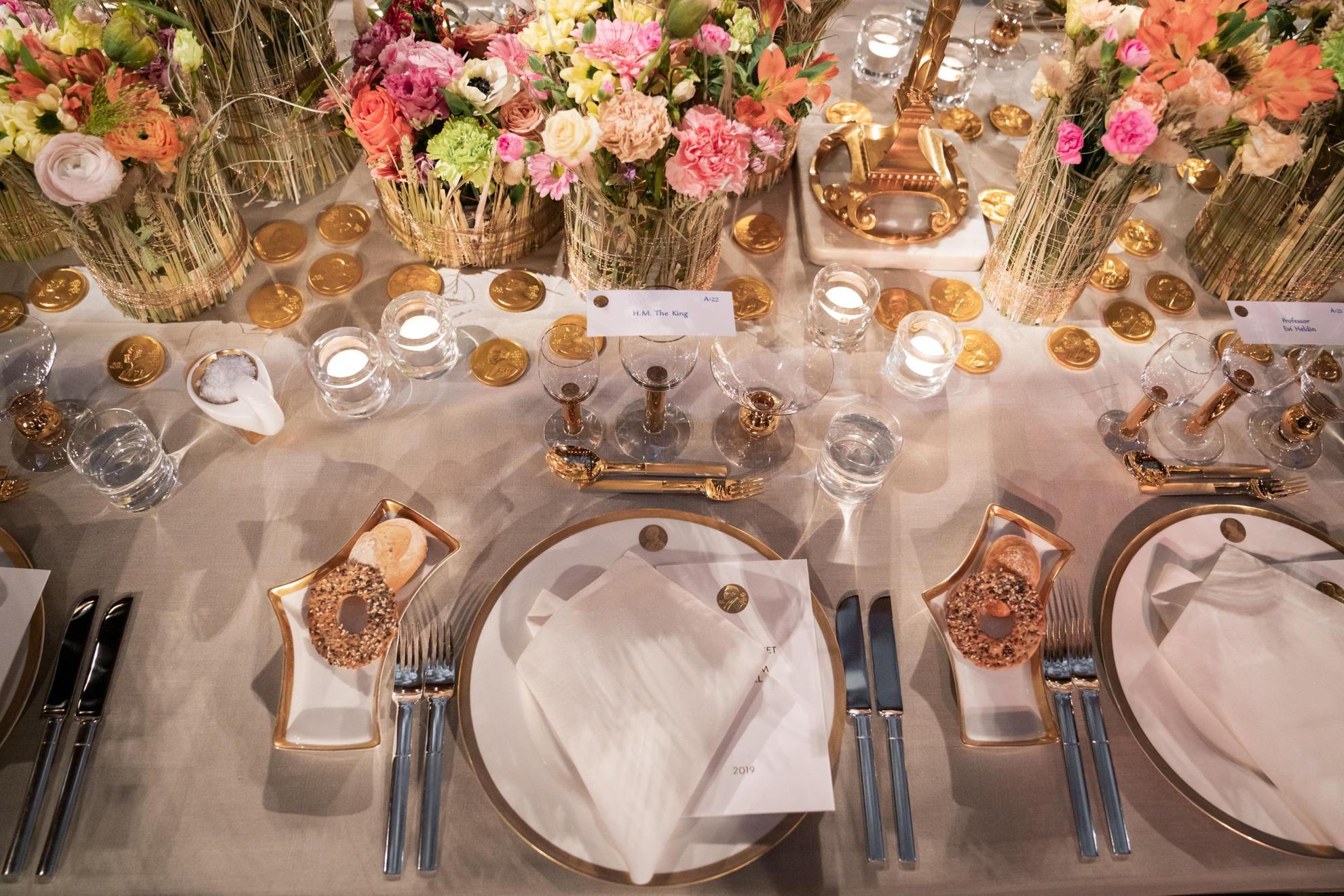 A tableware in white with golden details is set on a table with a white tablecloth. In the middle of the table are colorful flowers in vases.