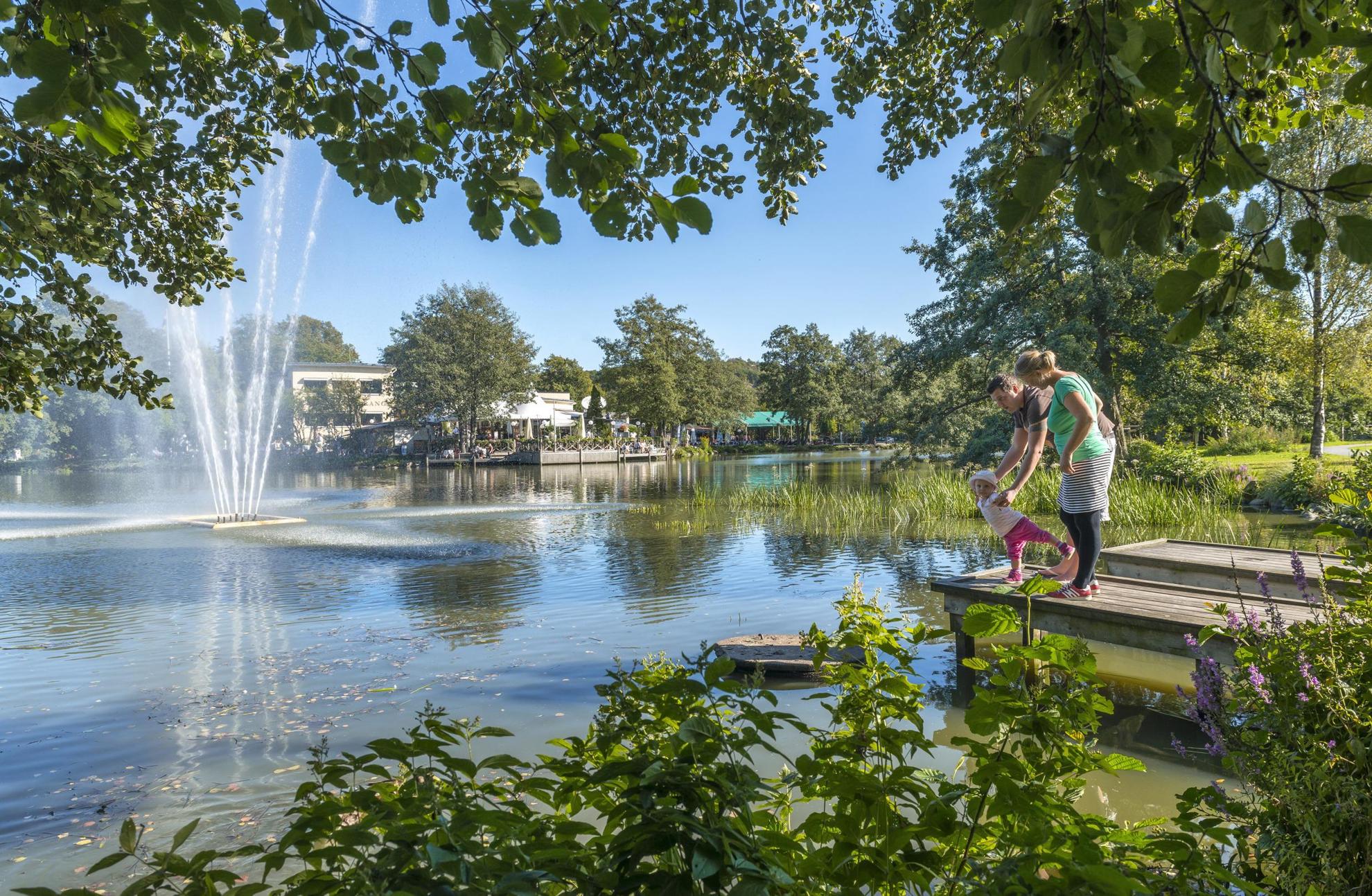 A couple with a small child standing on a jetty next to a pond with a fountain, surrounded by greenery in the park Slottsskogen. In the background you see an outdoor restaurant.