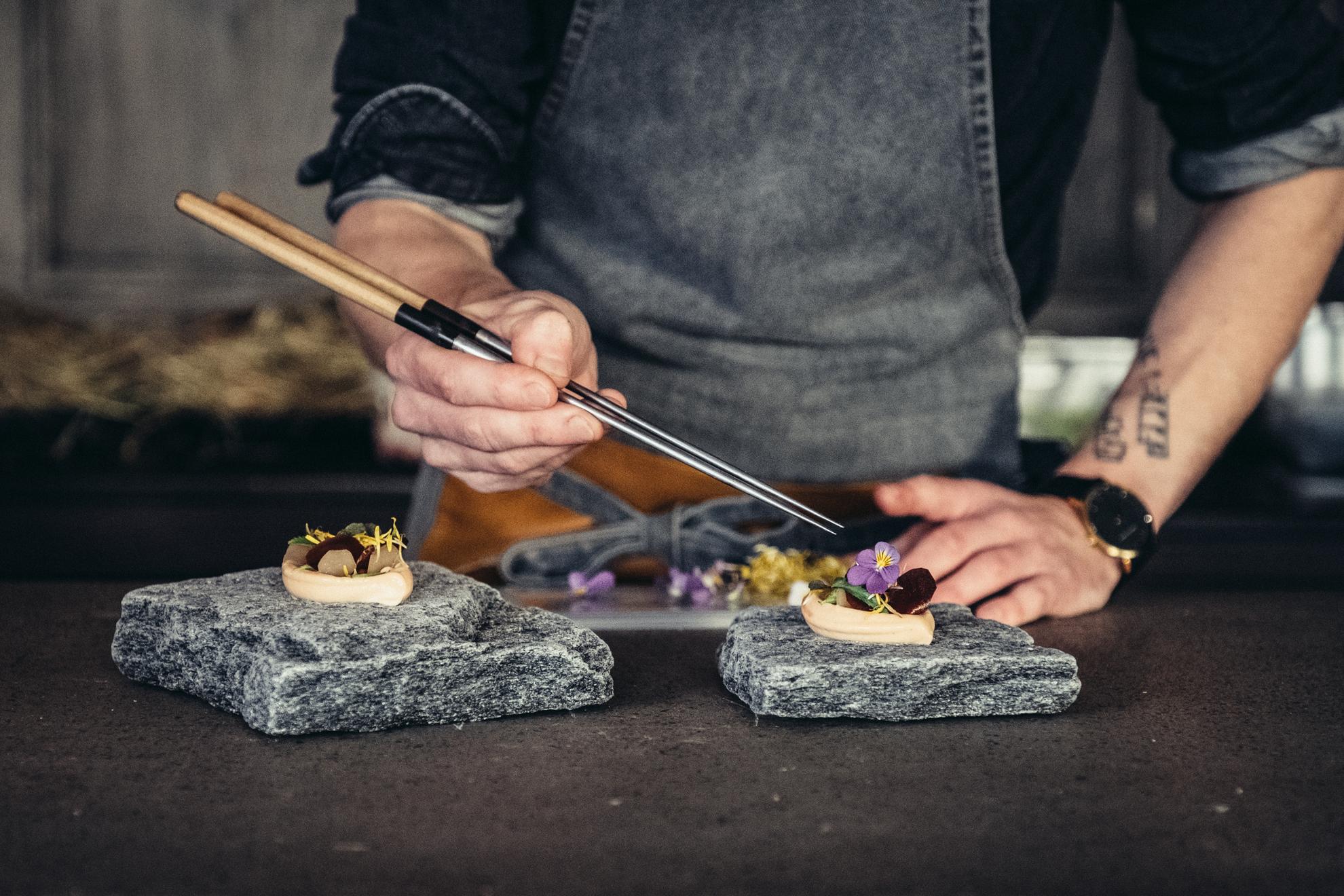 A person is plating food with a long tweezer. It's served on granite rocks and decorated with edible flowers.