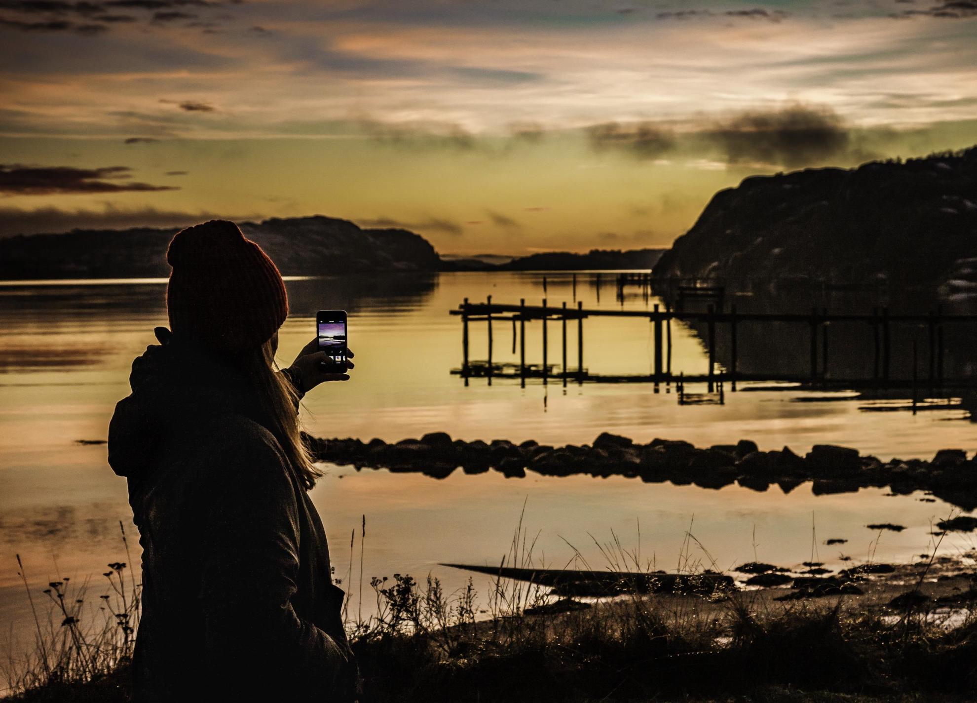 A woman is holding up a smartphone to a sunset by the water on the west coast of Sweden.