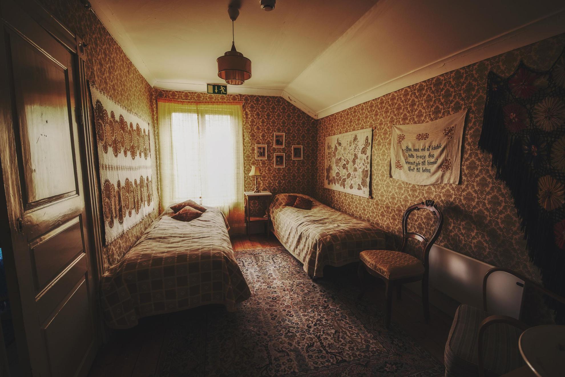 A room with decorative wallpaper, paintings, a rug, a chair and two single beds.