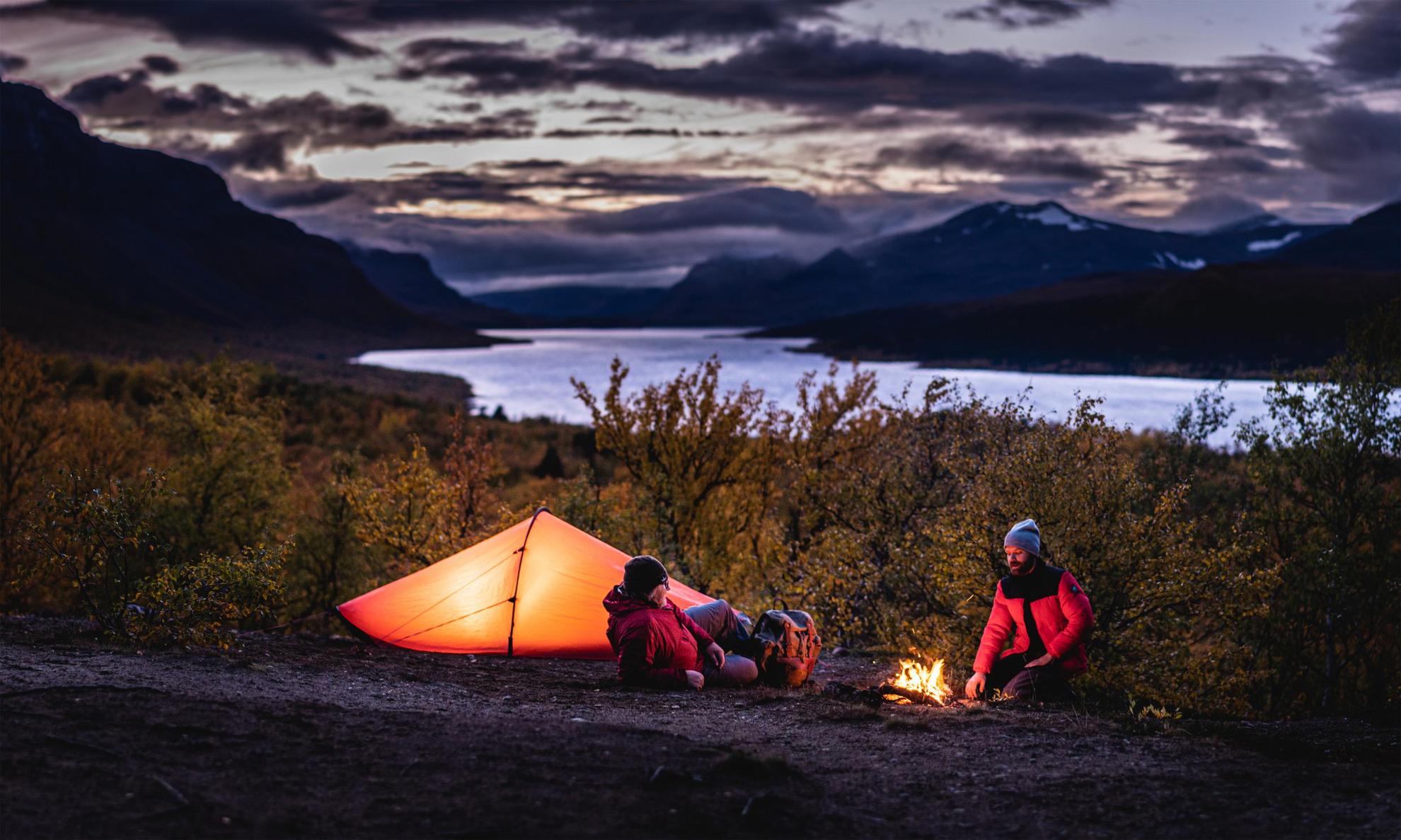 Two people sitting next to a tent and a camp fire. A river and mountains are in the background.