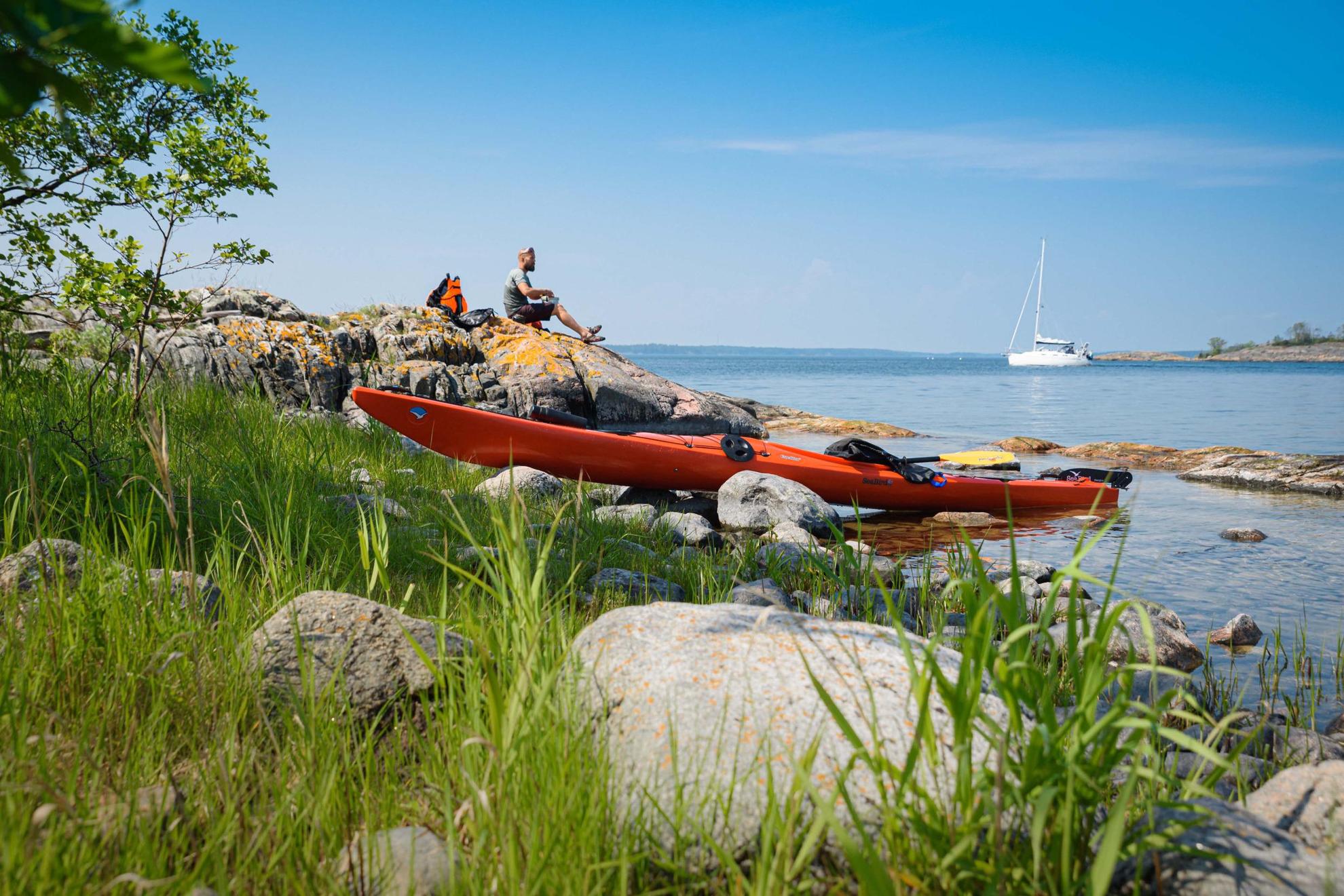 An orange kayak lies on the side of the water while a man enjoys a lunch on the cliffs. A white sailboat is in the background.