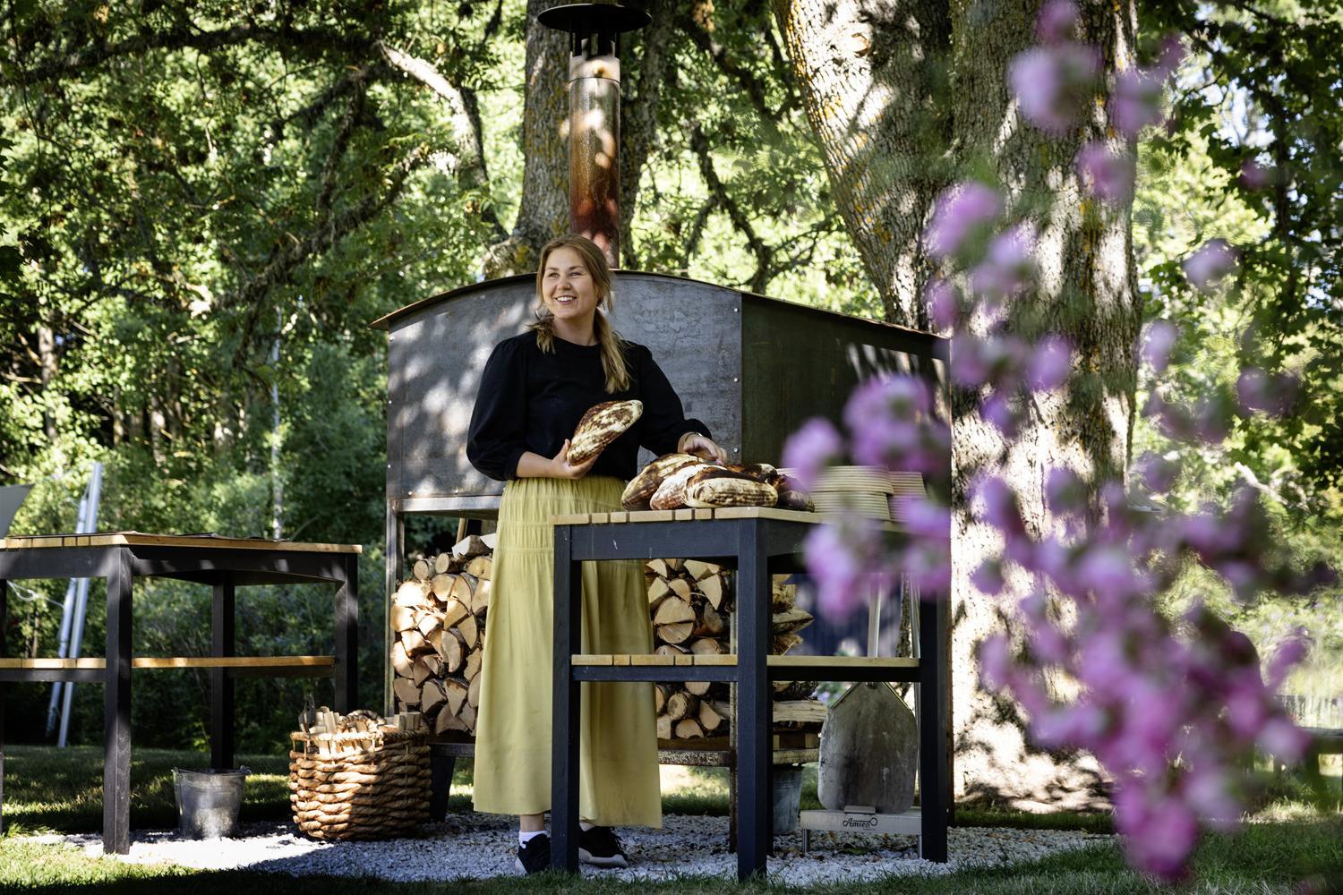 A woman is standing by a wood fired oven, holding bread, in a garden during summer.