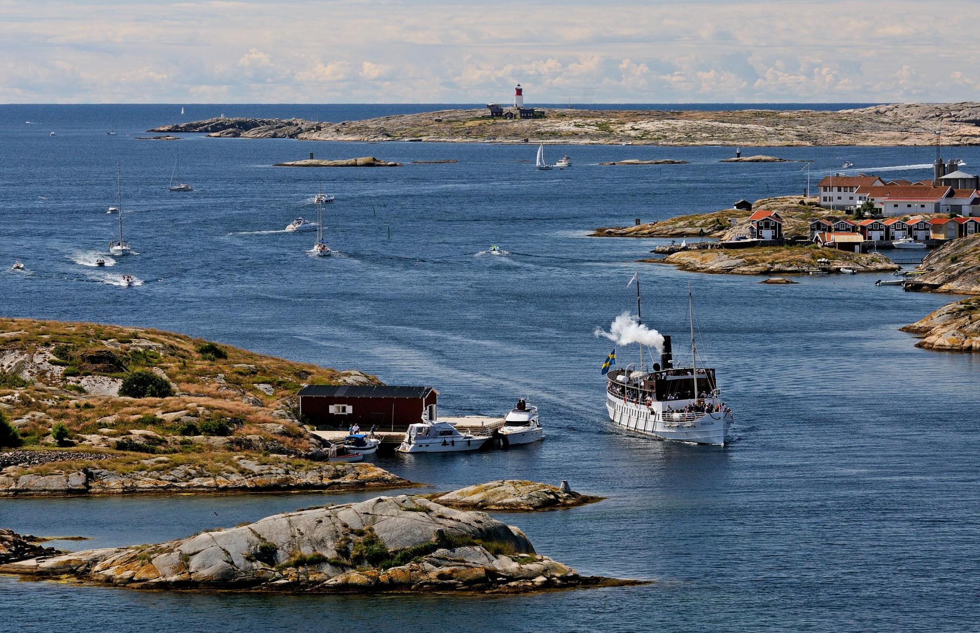 A steam boat and other sailing boats cruise between smaller islands in the archipelago. A bigger island in the background has a lighthouse.