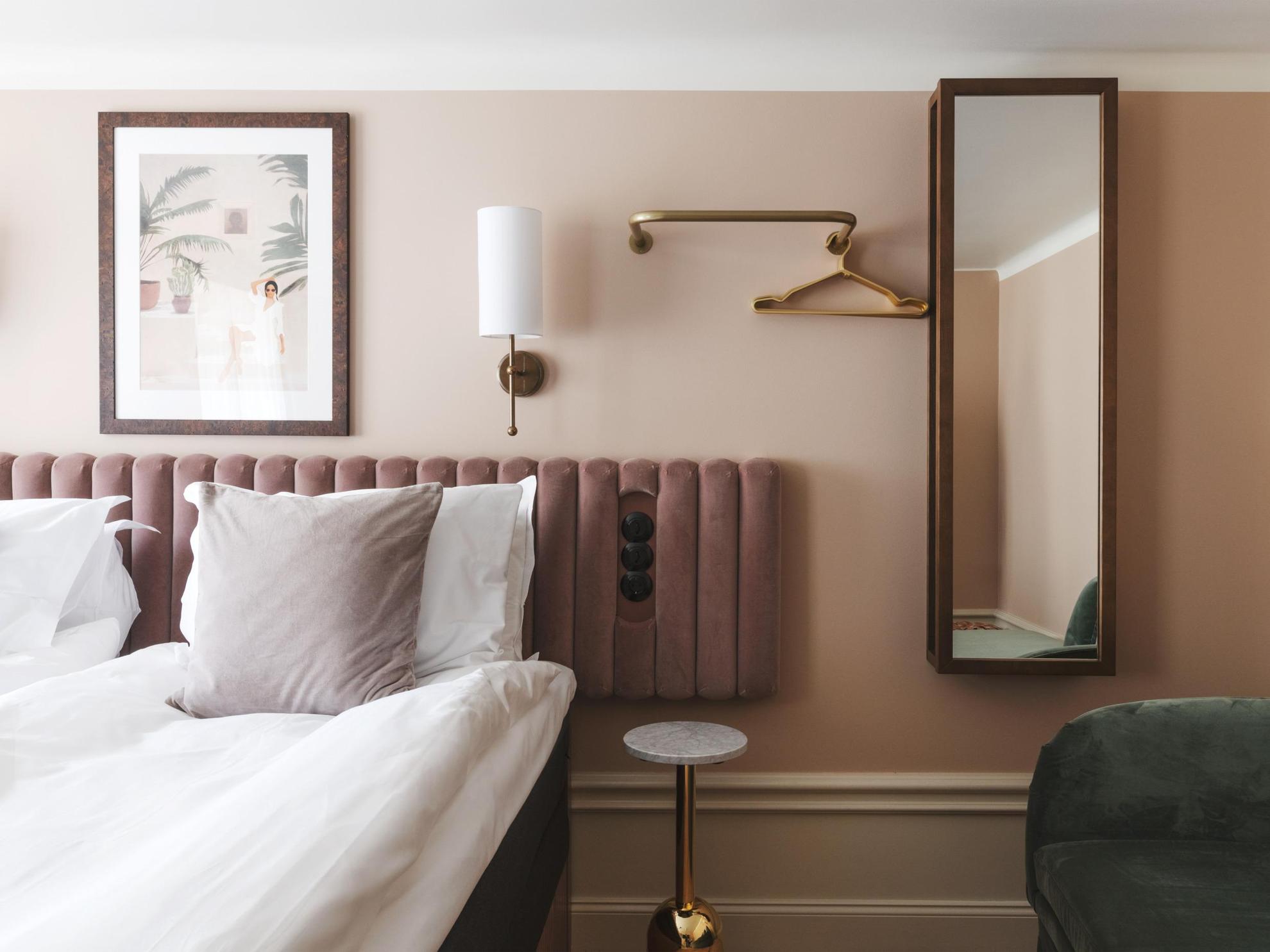A hotel room with beige-painted walls and a white ceiling. A brass wall lamp, a painting, a mirror and a coat hanger decorate the walls next to a double bed.