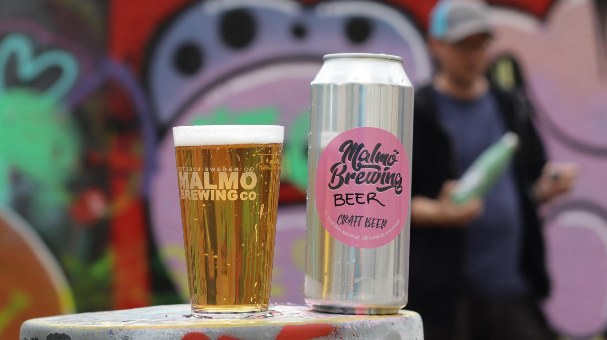 A beer can and a glass filled with beer. Out of focus in the background is a person and a graffiti wall.