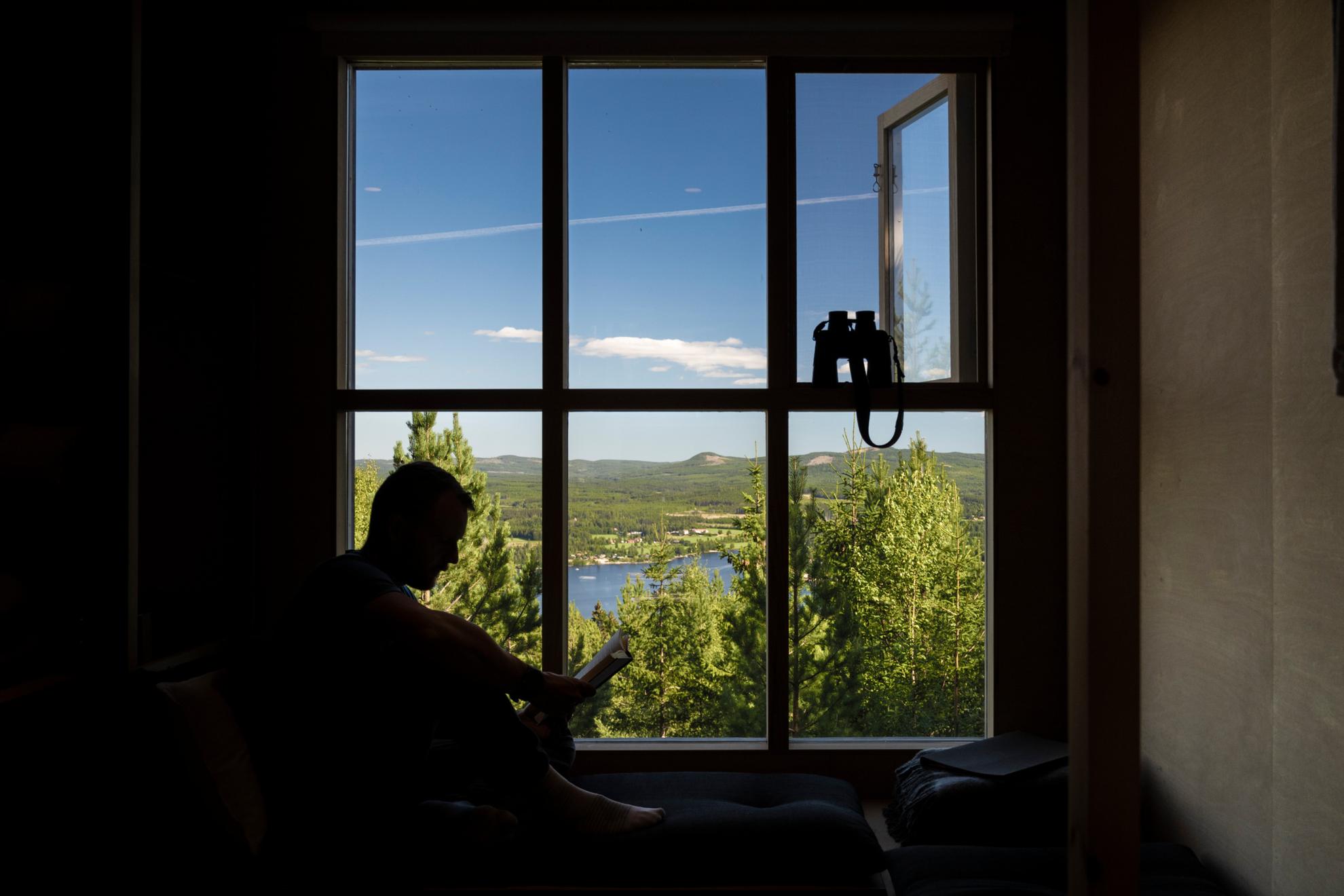 Silhouette of a man reading a book against a large window looking out over the blue sky and the mountains above in Hälsingland, with a pair of binoculars at the ready.