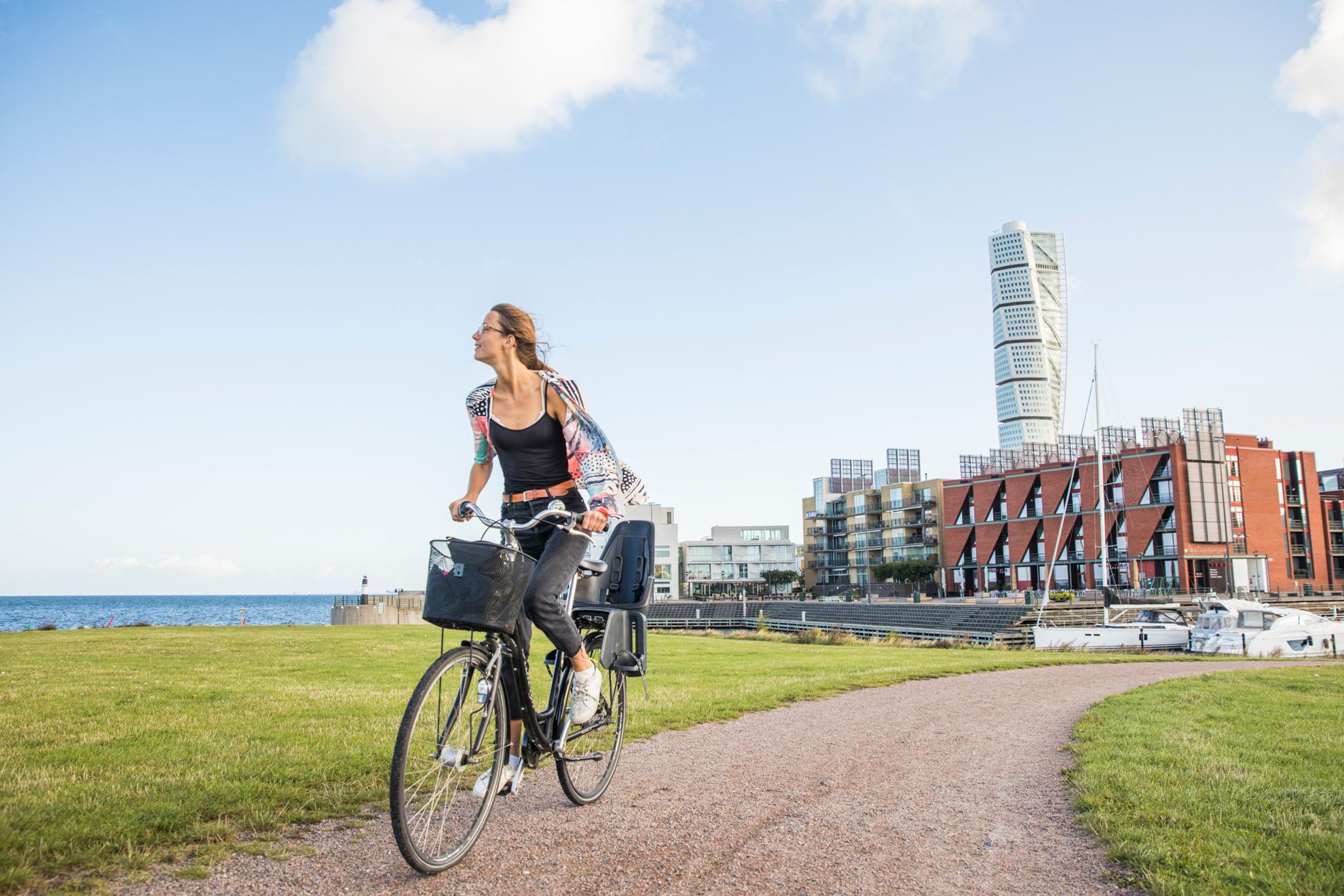 A woman ride a bike on a walkway, behind here you see a residential area by the sea and in the middle a skyscraper.